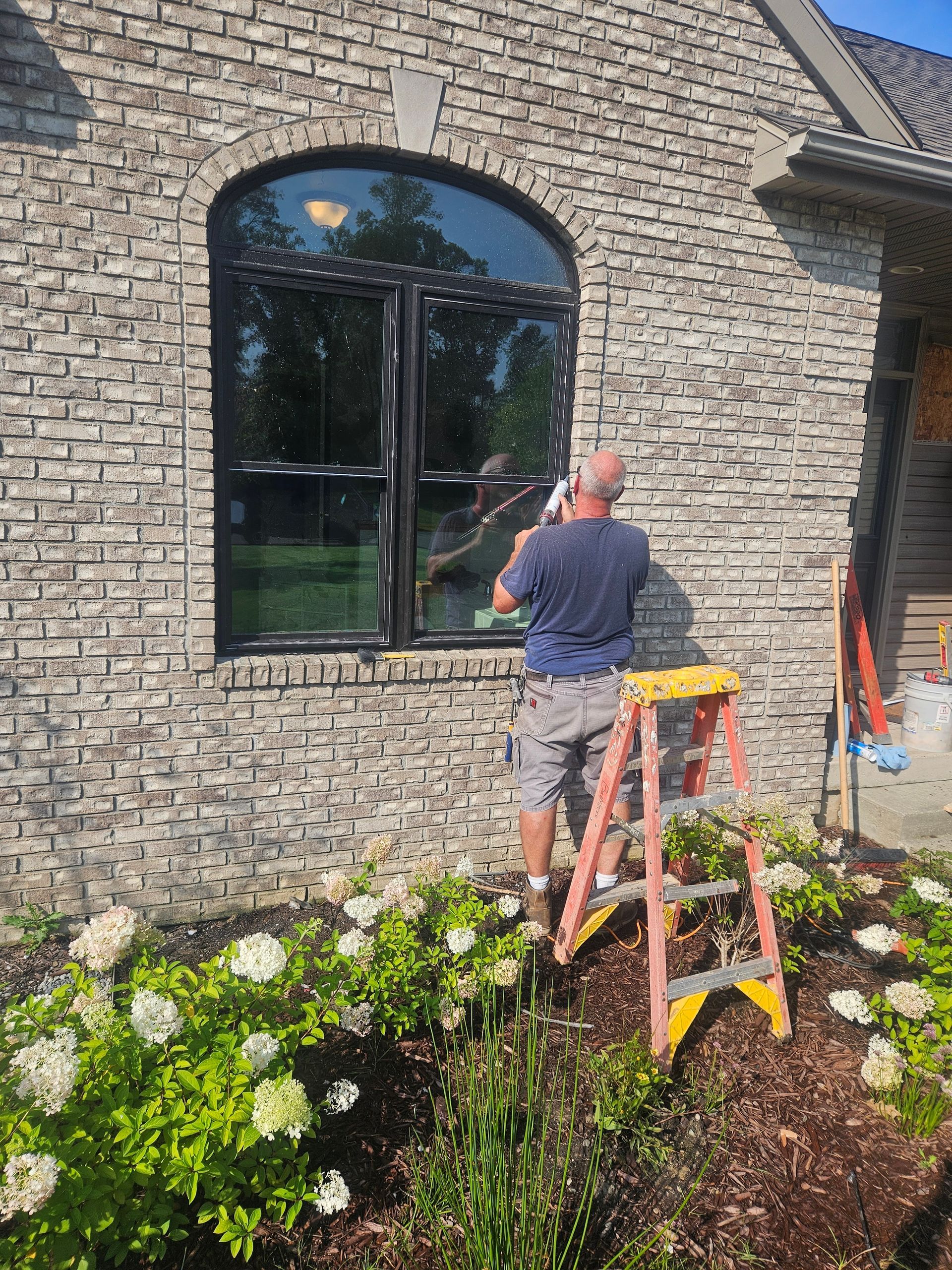 A man is standing on a ladder cleaning a window on the side of a brick building.
