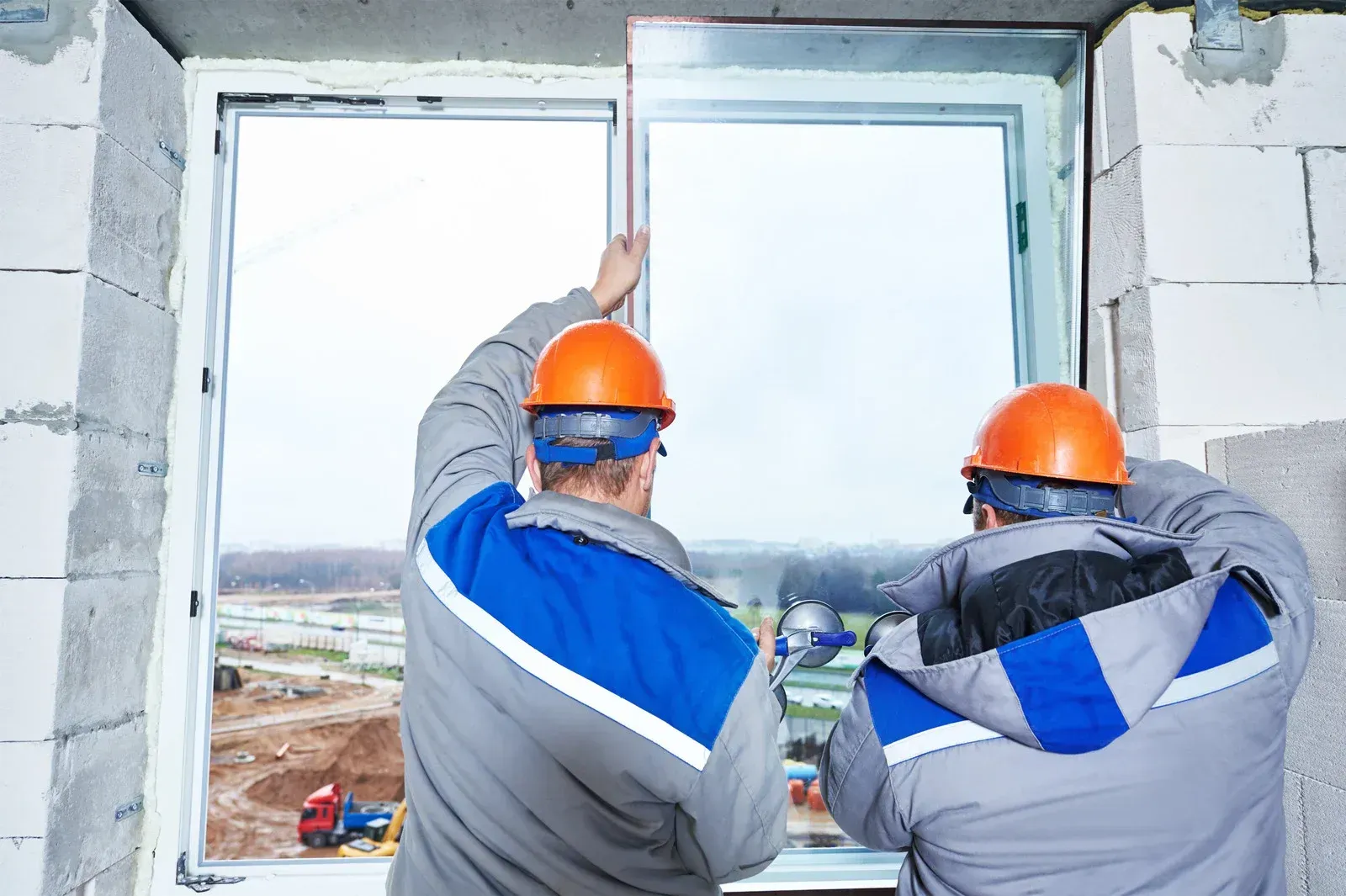 Two construction workers installing a window, wearing orange hard hats and blue and gray jackets.