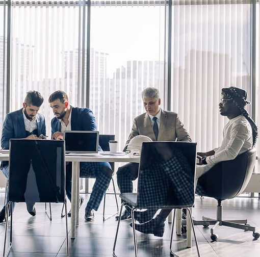 Business meeting at a table by a window; four people in suits review documents and use laptops.
