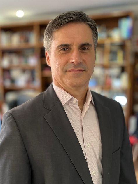 Um homem de terno e camisa branca está parado em frente a uma estante de livros.