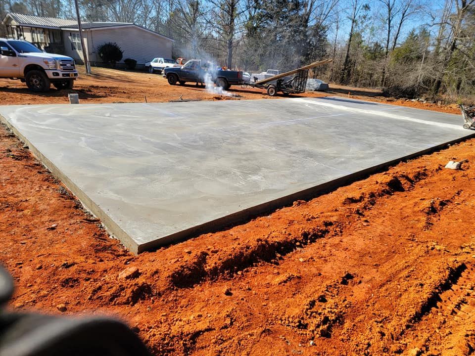 Freshly poured concrete slab in a dirt yard with a pickup truck and structures in the background.