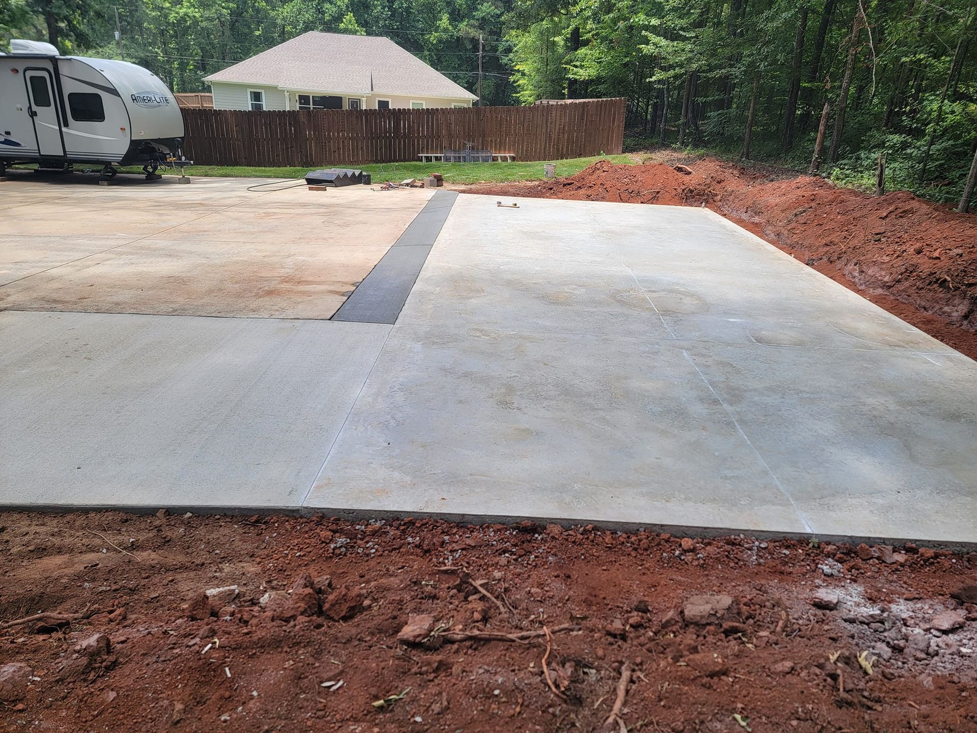 Concrete pad and driveway with camper in the background, surrounded by dirt and a wooden fence.
