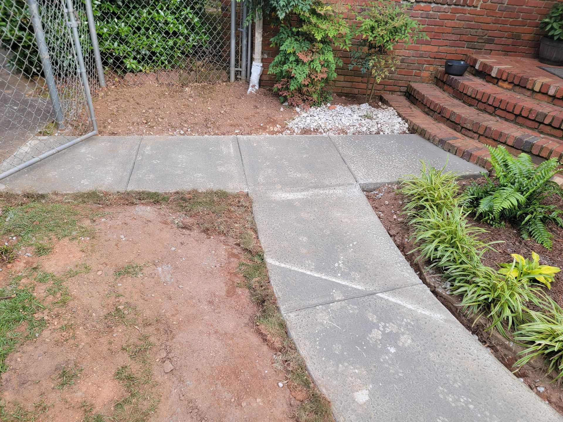 Concrete path leading to brick steps, edged by soil and plants.