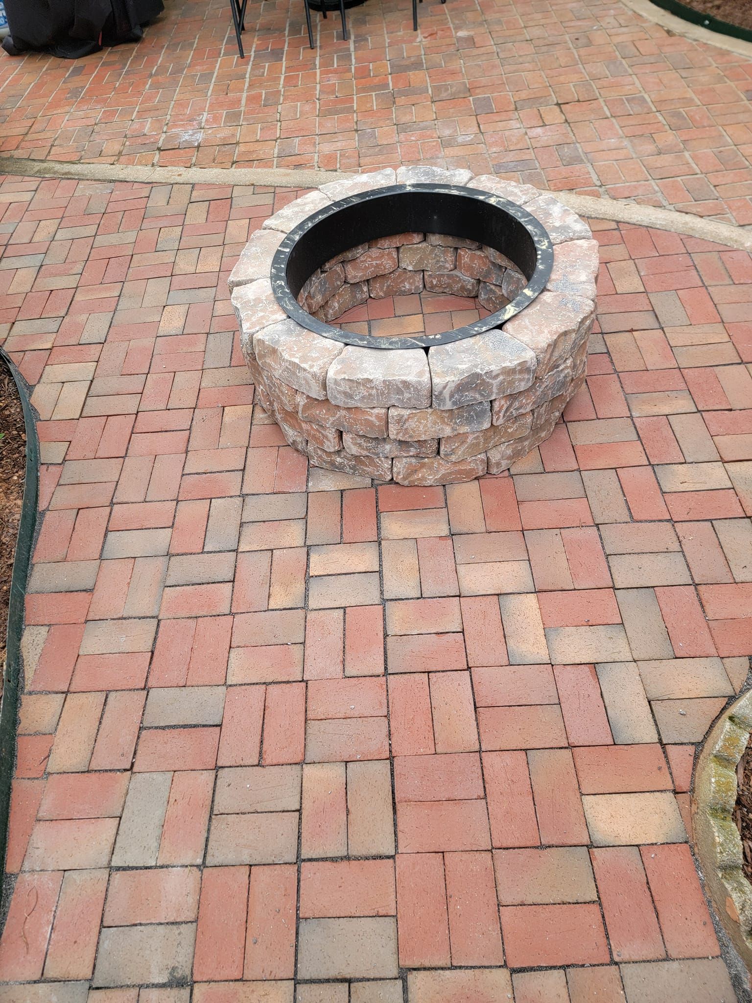 Fire pit on a brick patio. Beige stone fire pit with black metal interior, surrounded by red and brown bricks.