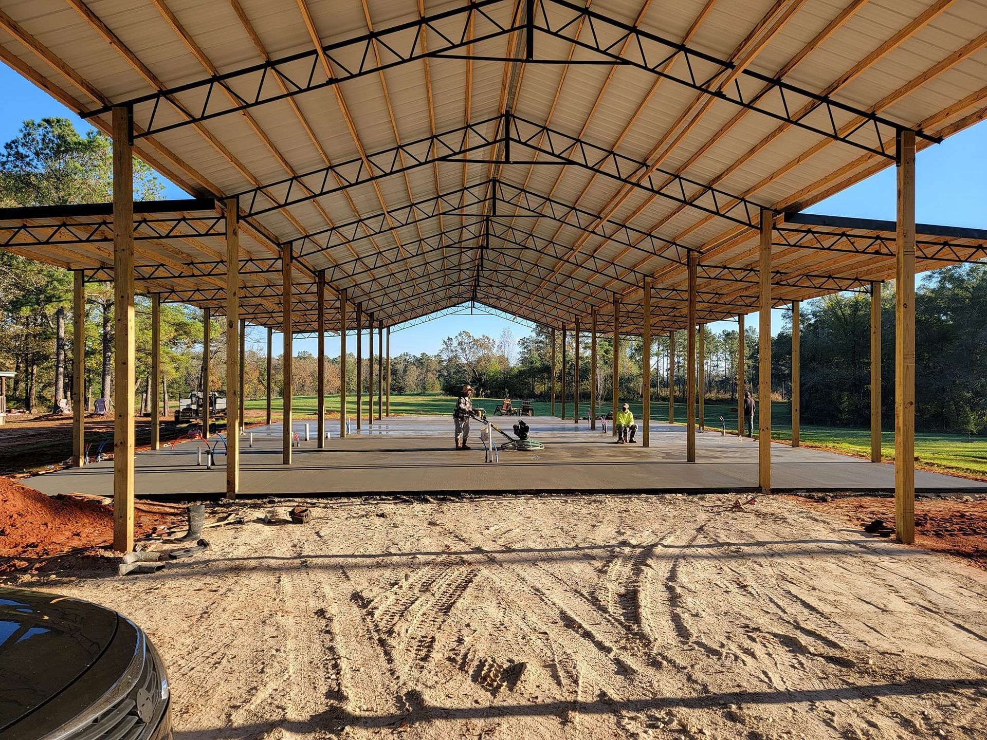 Open-air building under construction with concrete floor, wood supports, and metal roof in a rural setting.