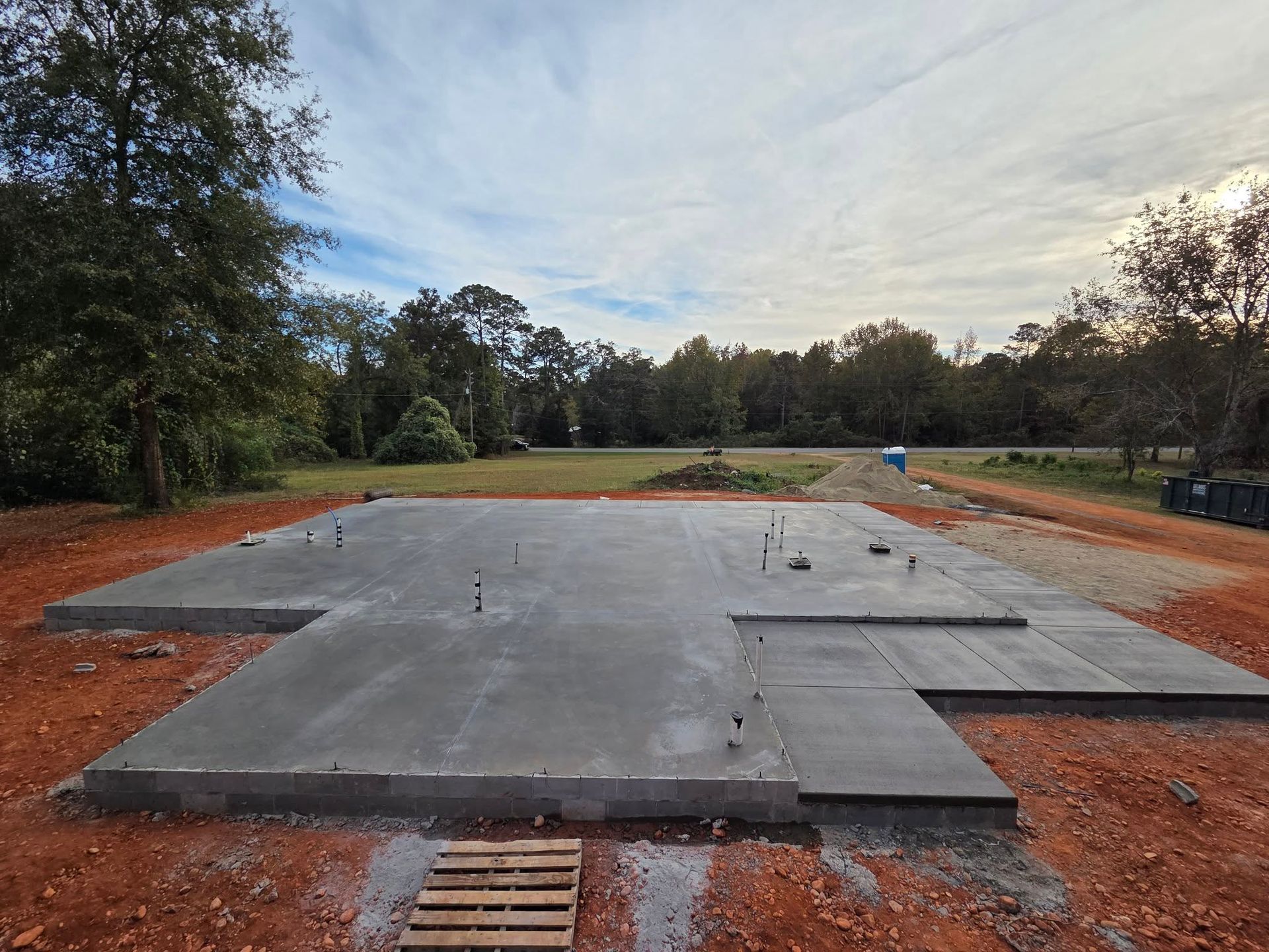 Newly poured concrete foundation in a rural setting, with plumbing fixtures and surrounding dirt.