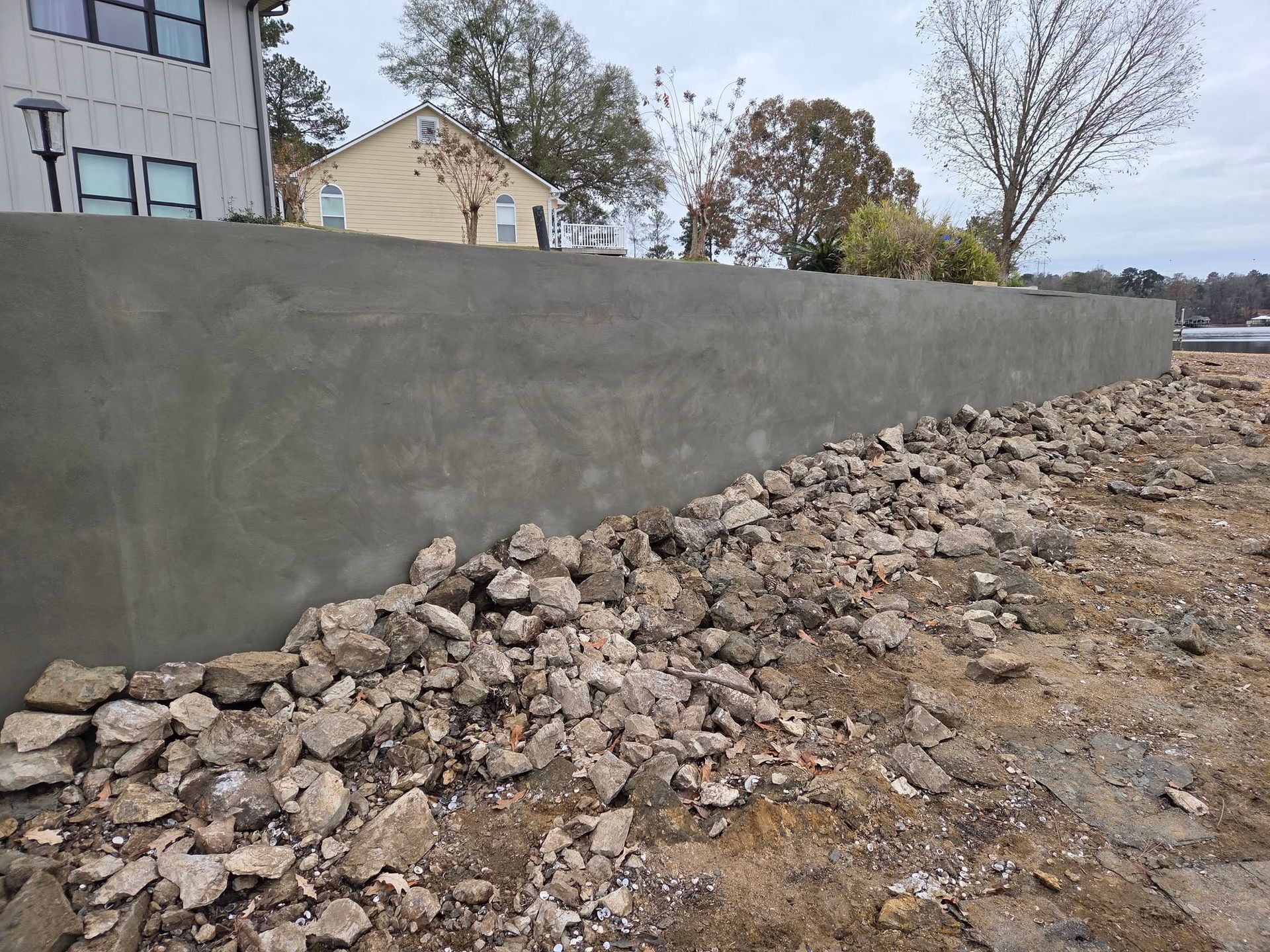 Concrete retaining wall, gray, with pile of rocks at its base. Houses and trees in background.