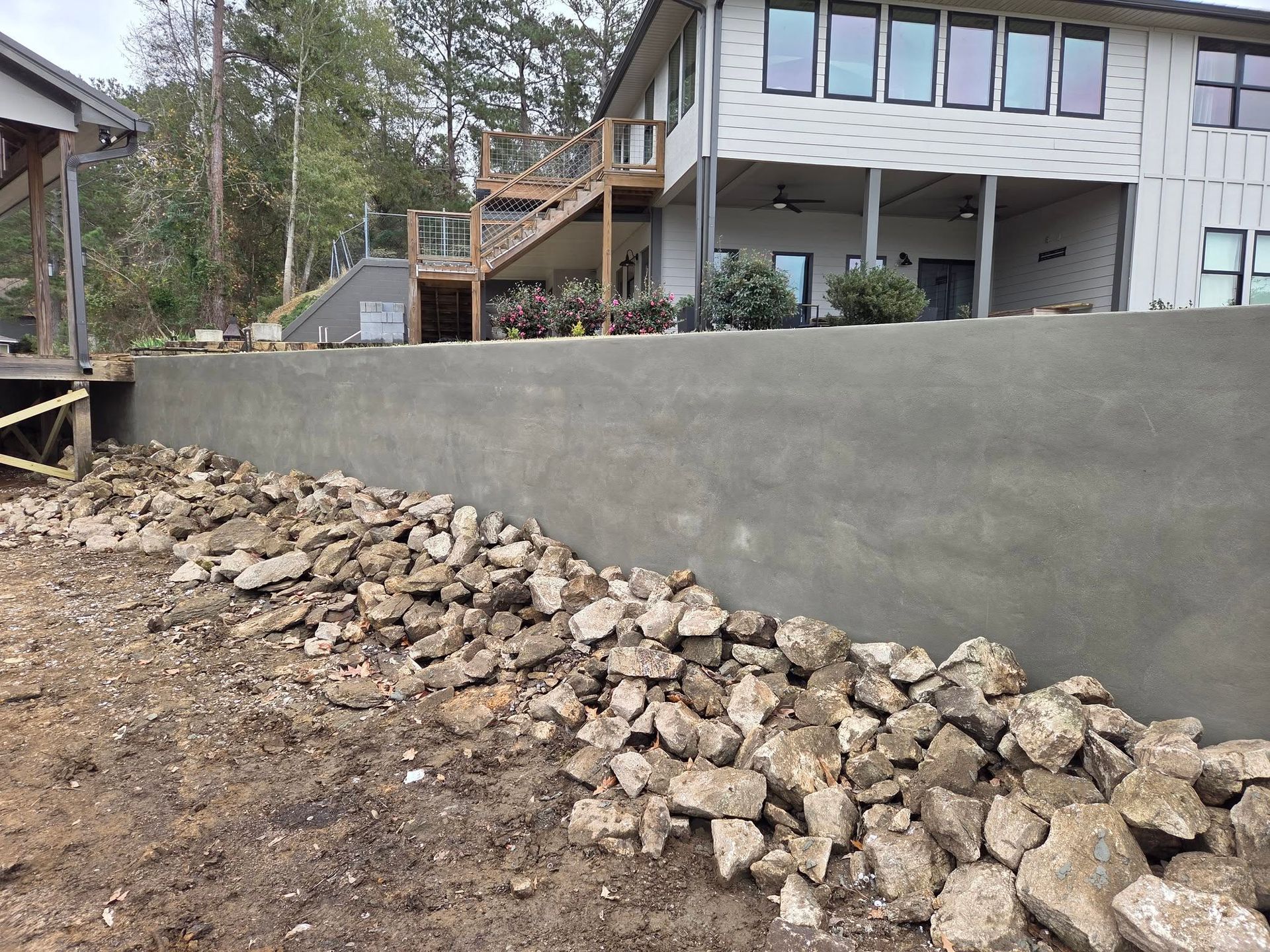 Gray retaining wall with rocks at the base, next to a two-story house with a deck.