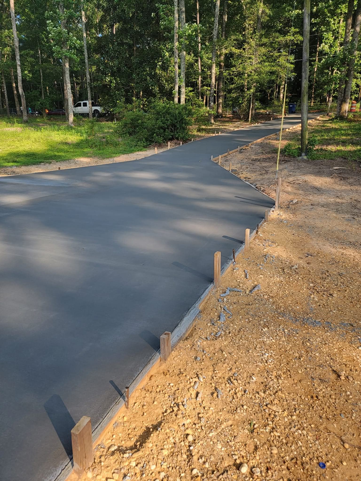Newly poured dark gray concrete driveway with wood formwork, gravel base, and trees in background.