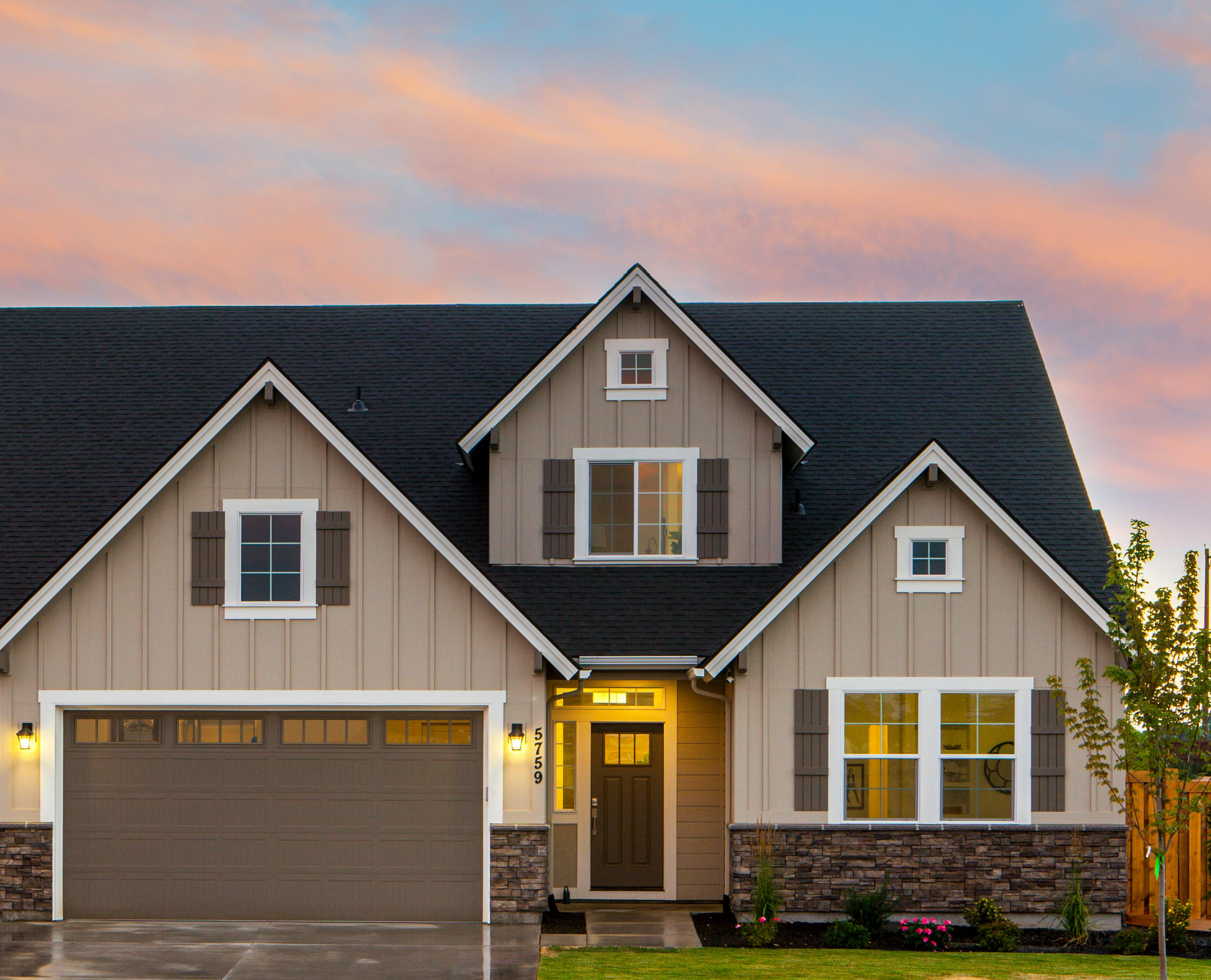 A two-story suburban house with beige vertical siding, a dark roof, a two-car garage, and stone accents at sunset.
