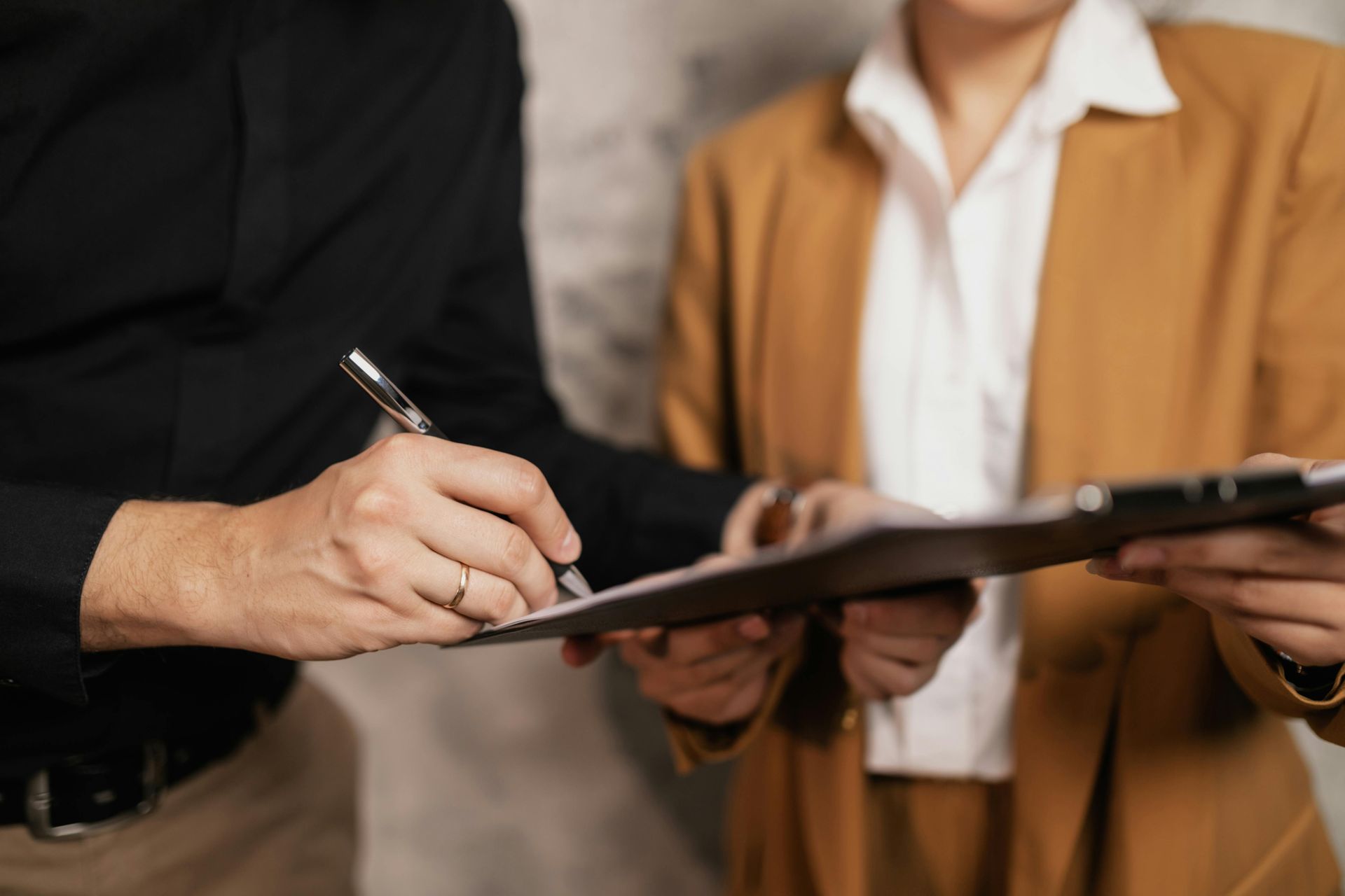 A person in a black shirt signs a document held by another person wearing a tan blazer and white collared shirt.
