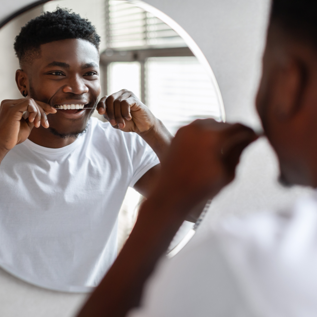 Man flossing his teeth in front of a mirror, smiling, wearing a white t-shirt.