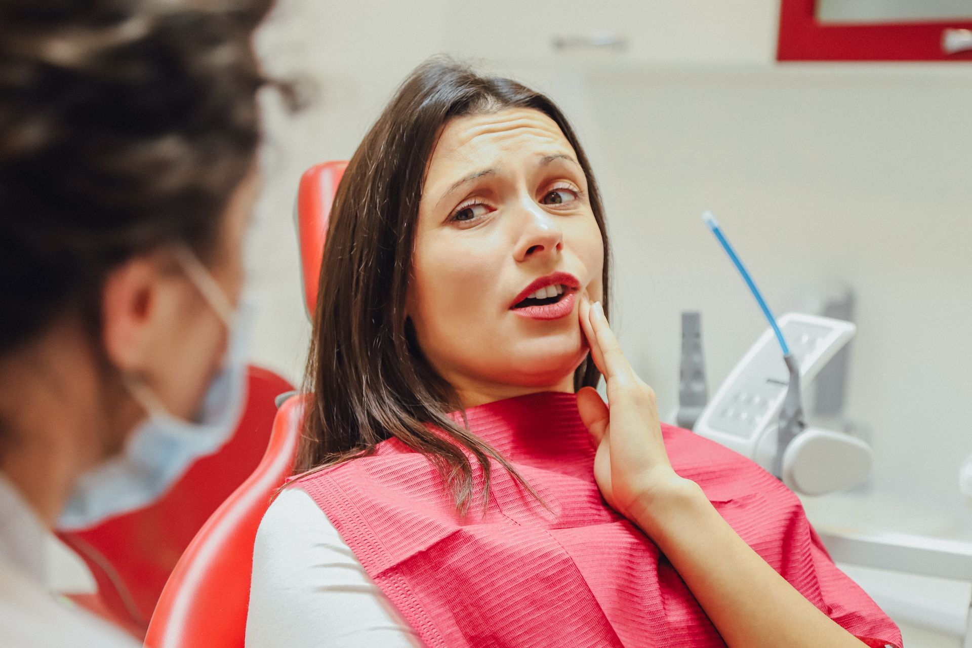 Woman in dental chair, hand on cheek, appears in pain, dentist with mask beside.