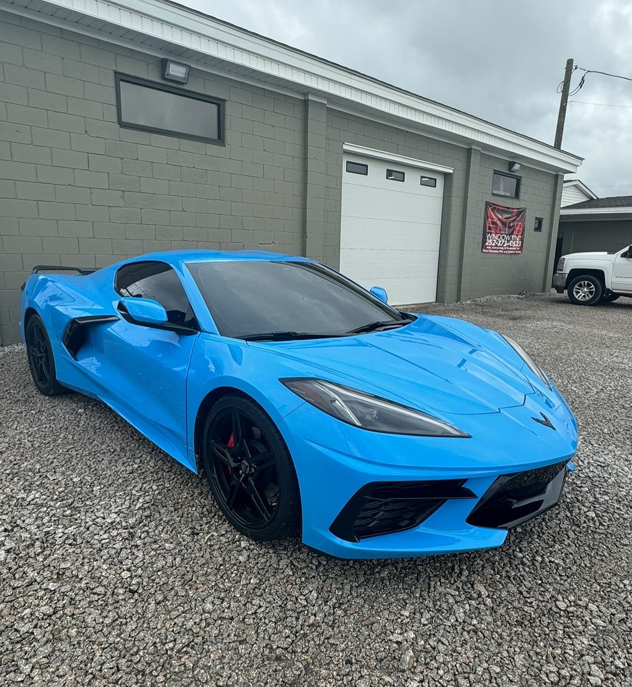 Bright blue Chevrolet Corvette parked on gravel in front of a gray building.