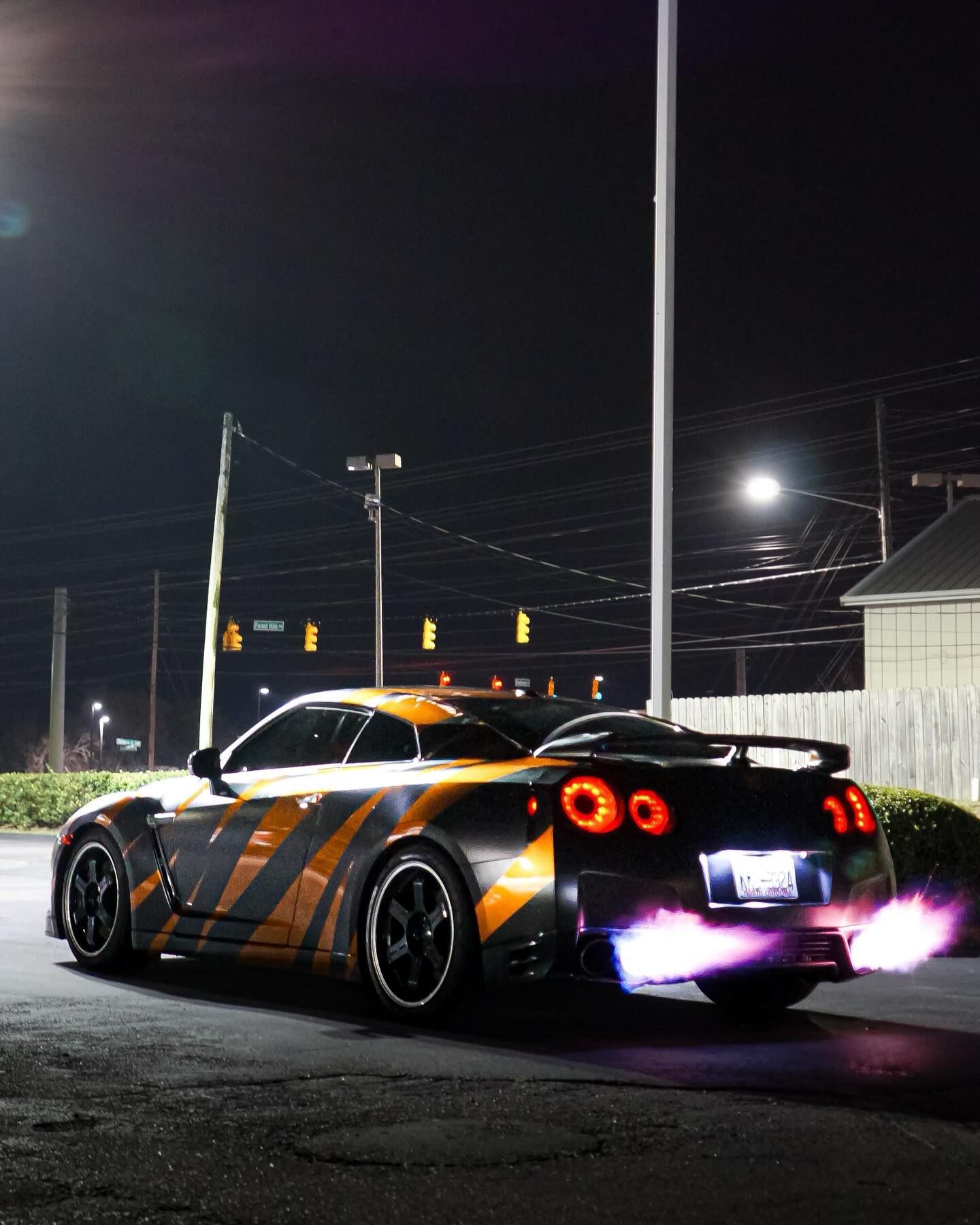 Nissan GT-R sports car with orange and black graphics at night, exhaust flames visible.