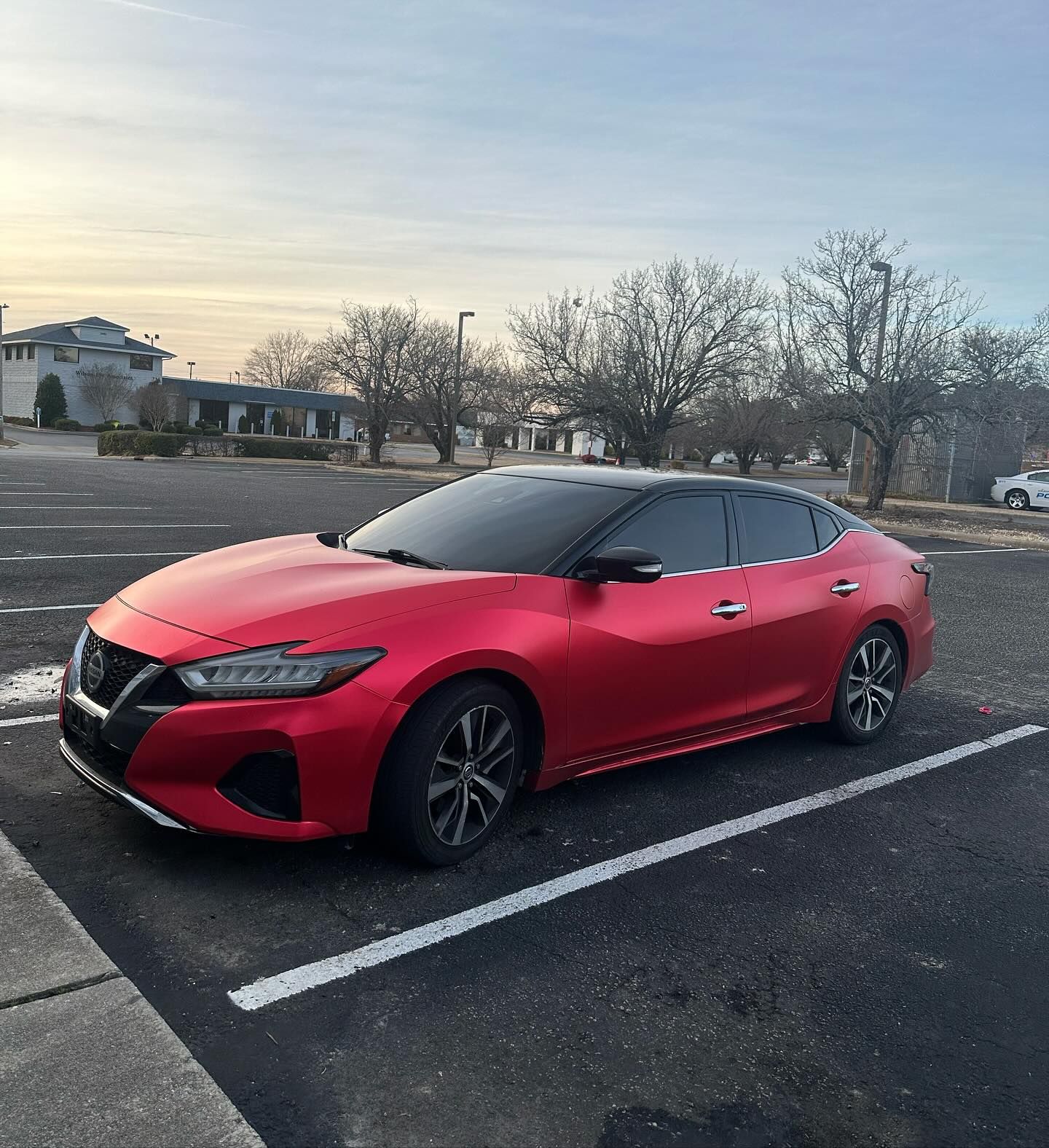 Red Nissan Maxima parked in a parking lot with a black roof. Evening sky in the background.