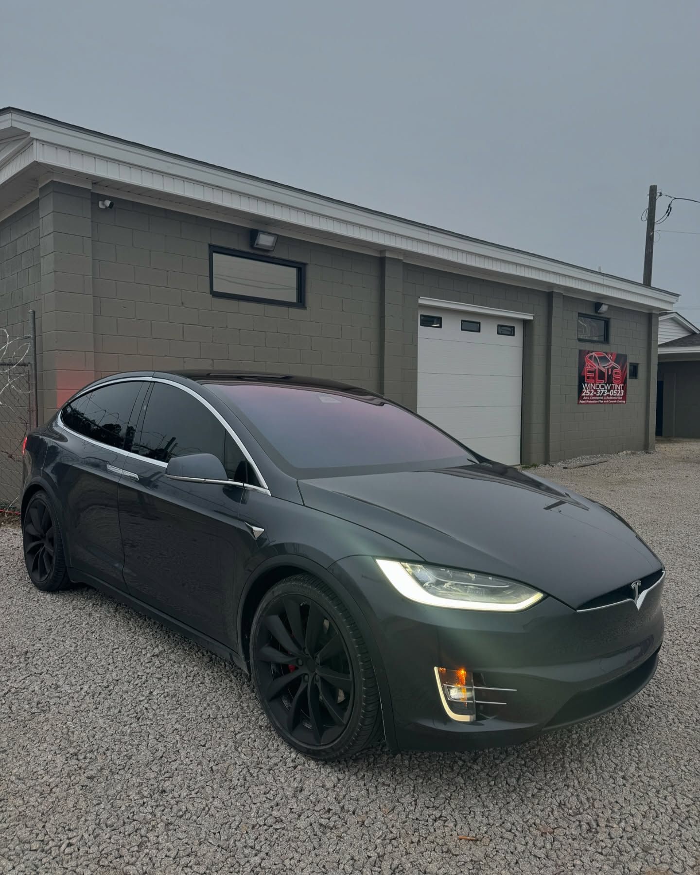 Dark gray Tesla Model X parked outside a gray building with a closed garage door.