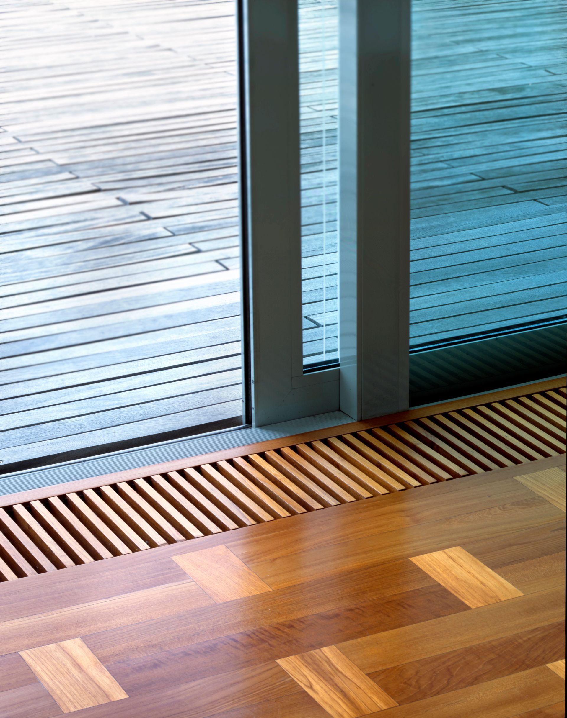 Una griglia di ventilazione a pavimento in legno scorre lungo una porta scorrevole in vetro che si apre su una terrazza in legno.