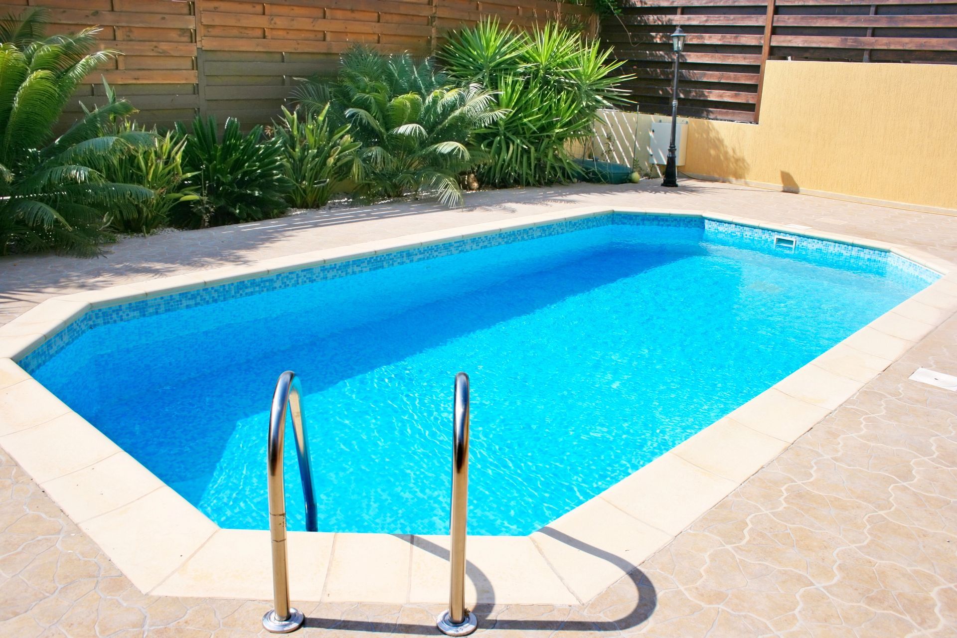 Una piscina rettangolare di un blu brillante, con ringhiere a scaletta in metallo, circondata da una pavimentazione in pietra e da una rigogliosa vegetazione.