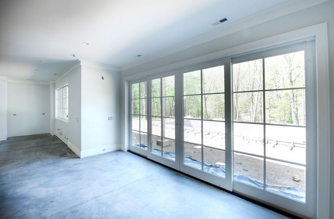 Una stanza spoglia con pavimento in cemento grigio, pareti bianche e un'ampia porta a vetri a quattro pannelli che si affaccia su una zona boschiva.