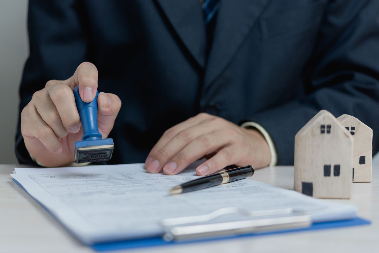 Person in suit stamps a document with a blue stamp near toy houses and a pen.