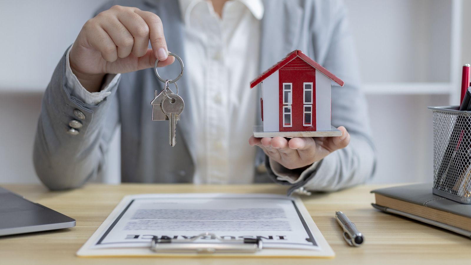 Person holding house keys and a model home over a desk with paperwork and a pen.