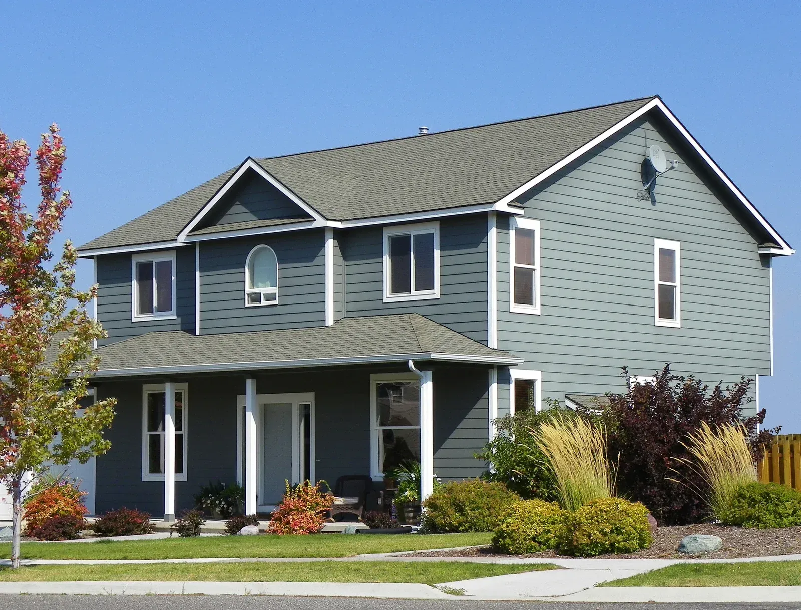 Two-story blue house with white trim, porch, and green roof, set in a sunny, landscaped yard.