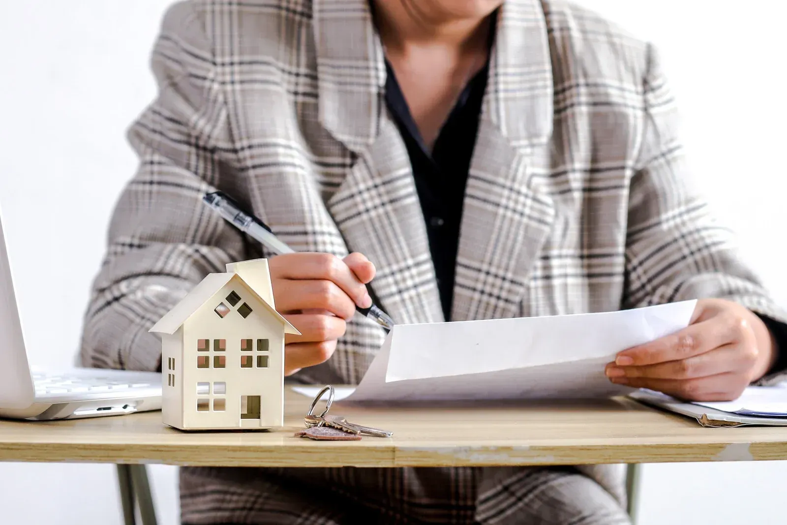 Person signing paperwork with a model house, keys, and laptop on a desk.