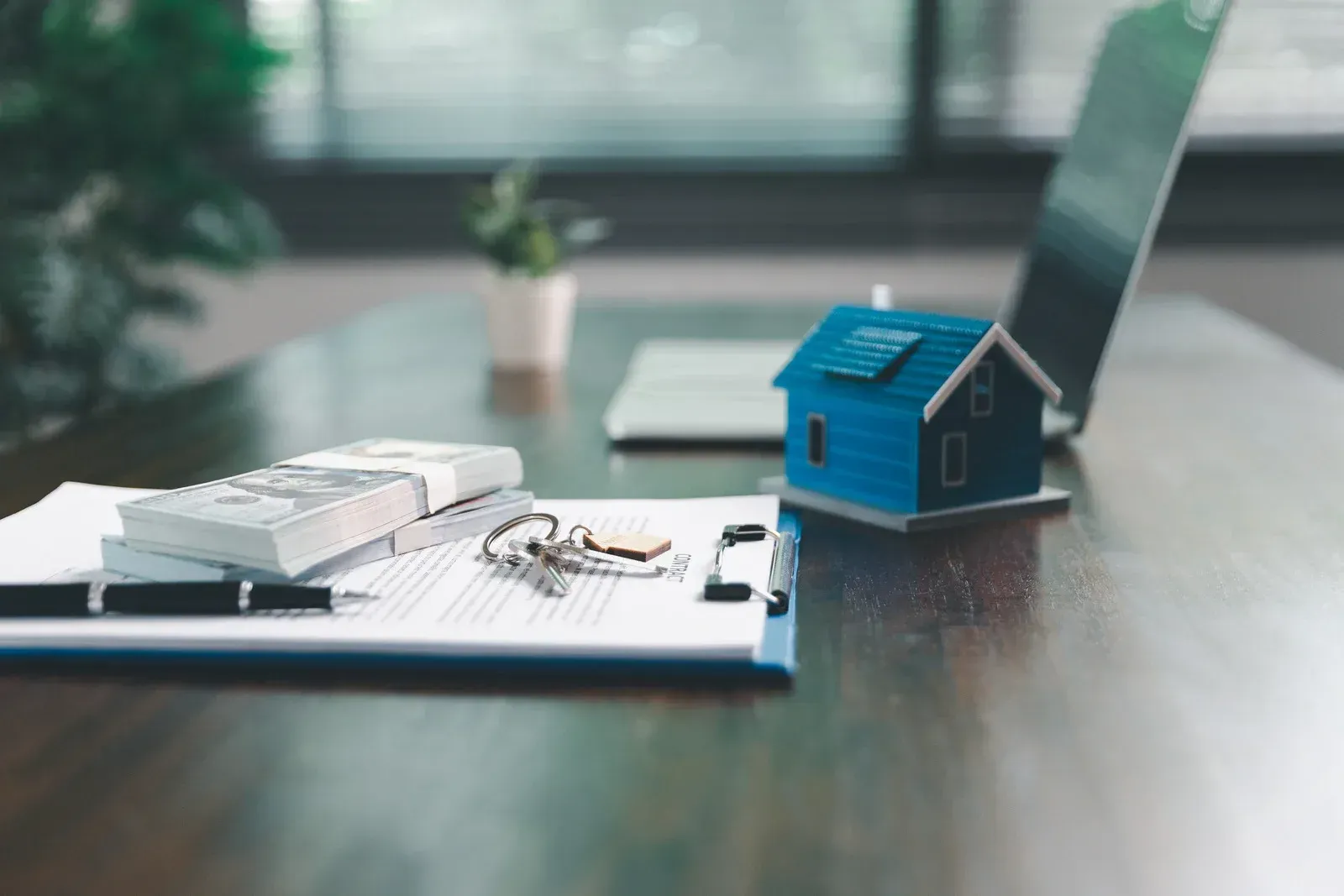 A desk with house model, cash, keys, pen, and laptop, suggesting a real estate transaction.