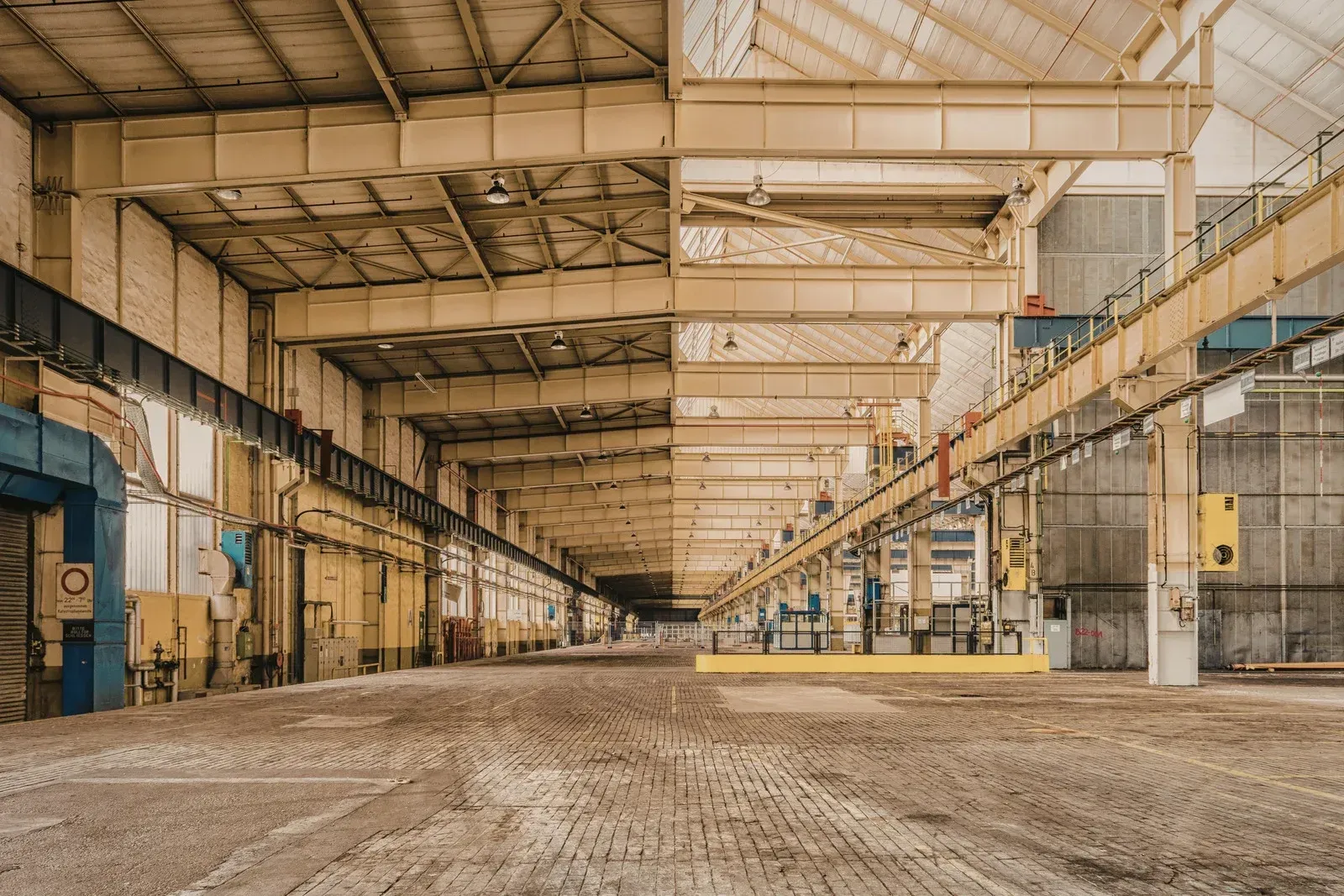 Empty industrial warehouse interior, brick floor, overhead crane, yellow and gray tones.