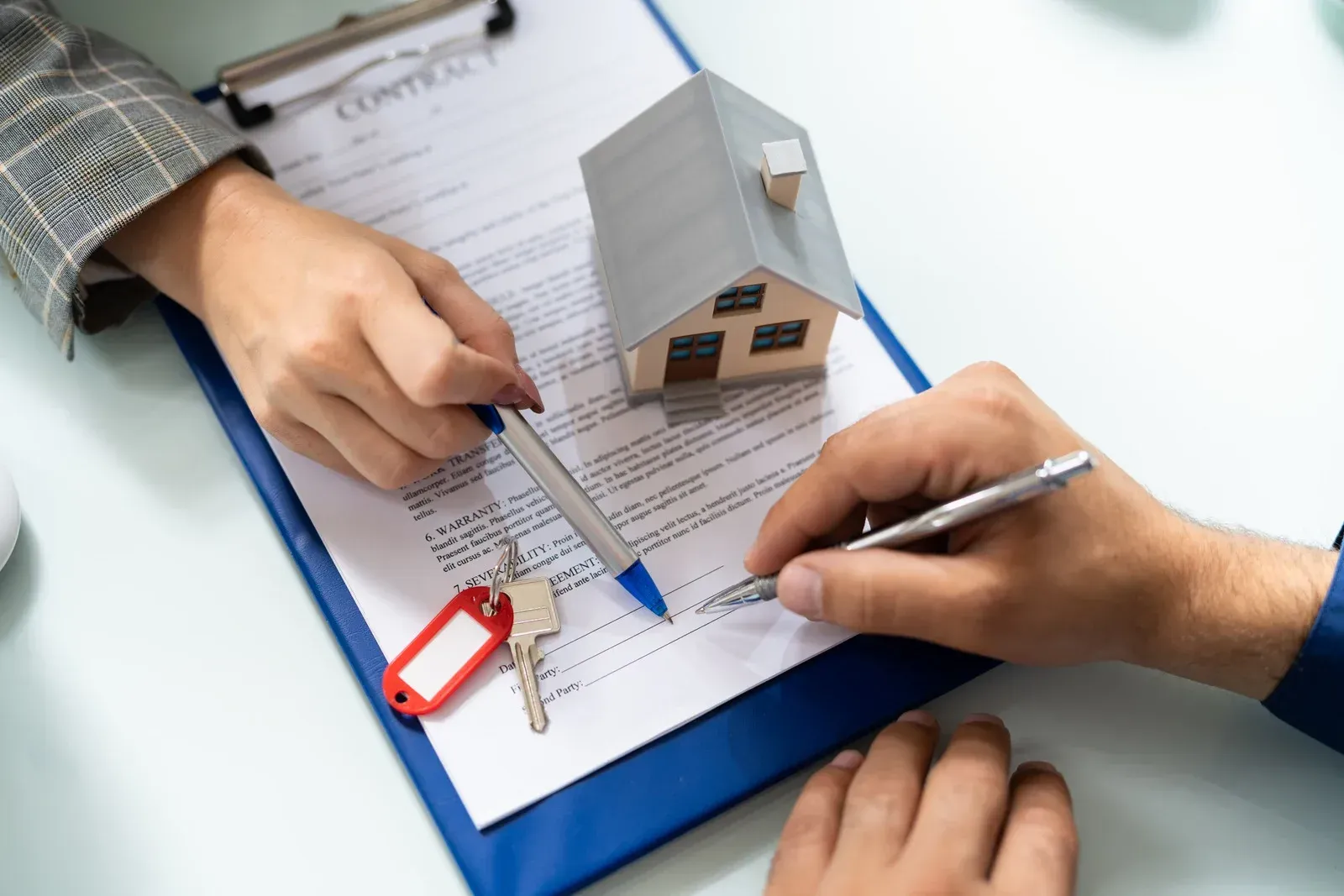 Hands signing a real estate contract with a miniature house and keys on a clipboard.