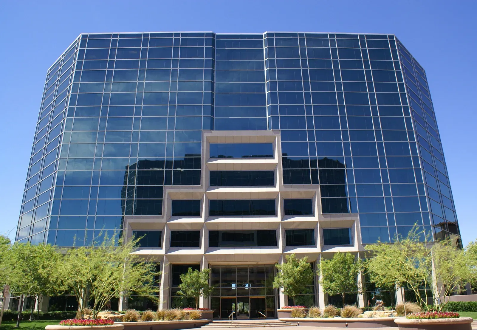 Glass-walled office building with a concrete entryway and landscaping under a blue sky.