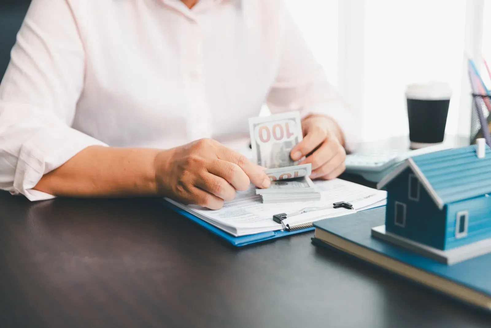 Person counting money over documents, miniature house and coffee cup on desk.