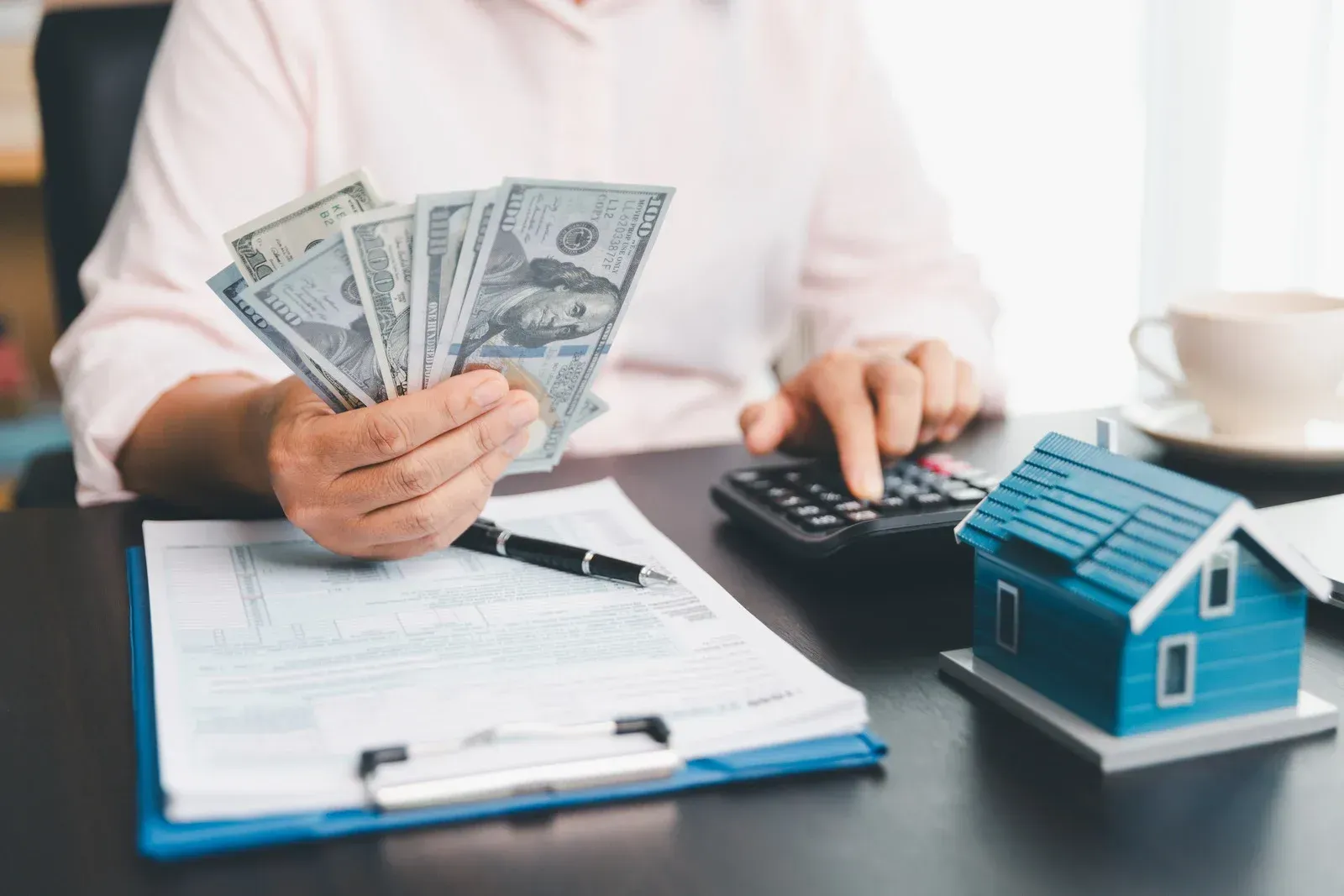 Person calculating finances, holding cash, with a toy house and paperwork on a desk.
