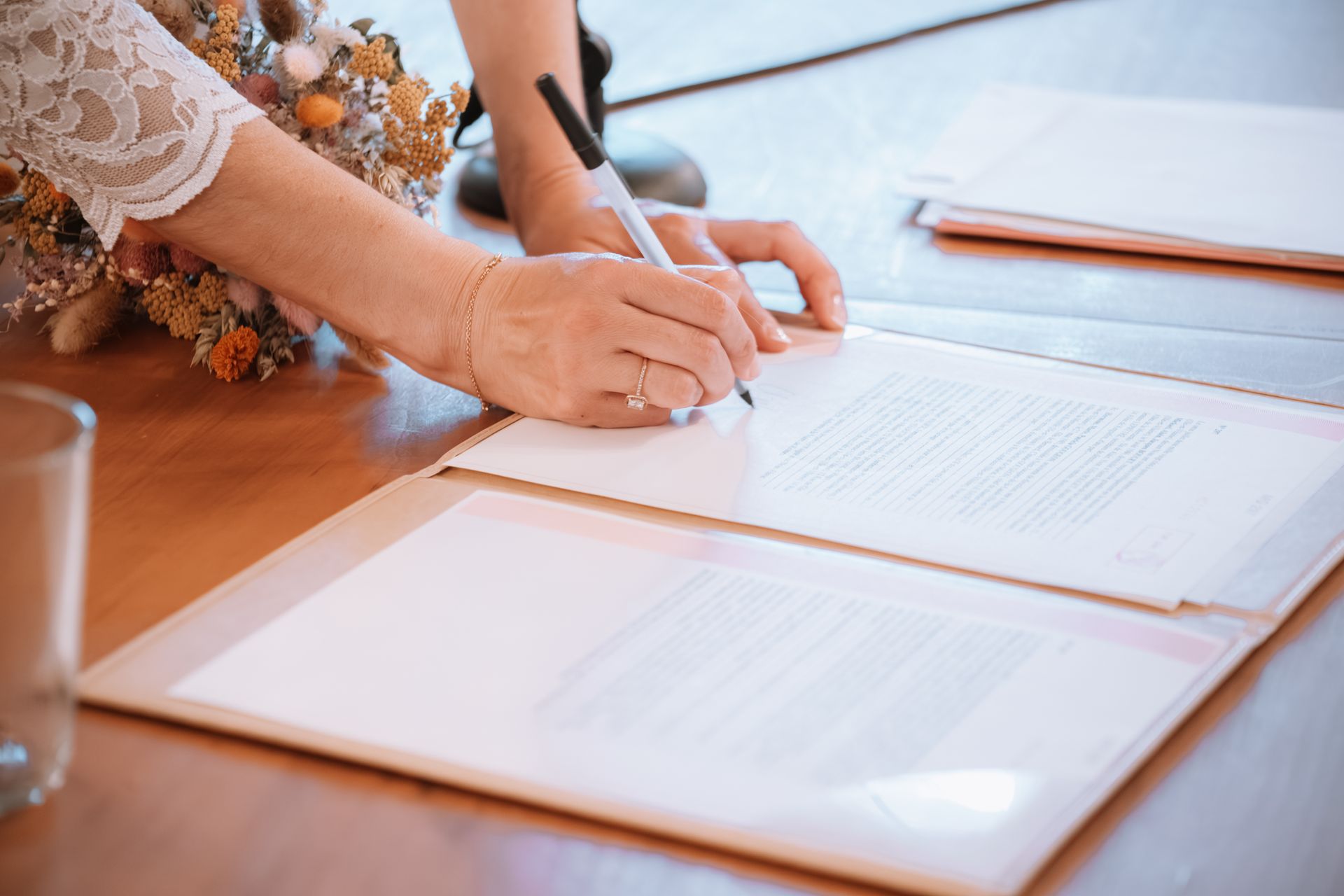 Person signing a document on a wooden table. A pen, flowers, and a glass are nearby.