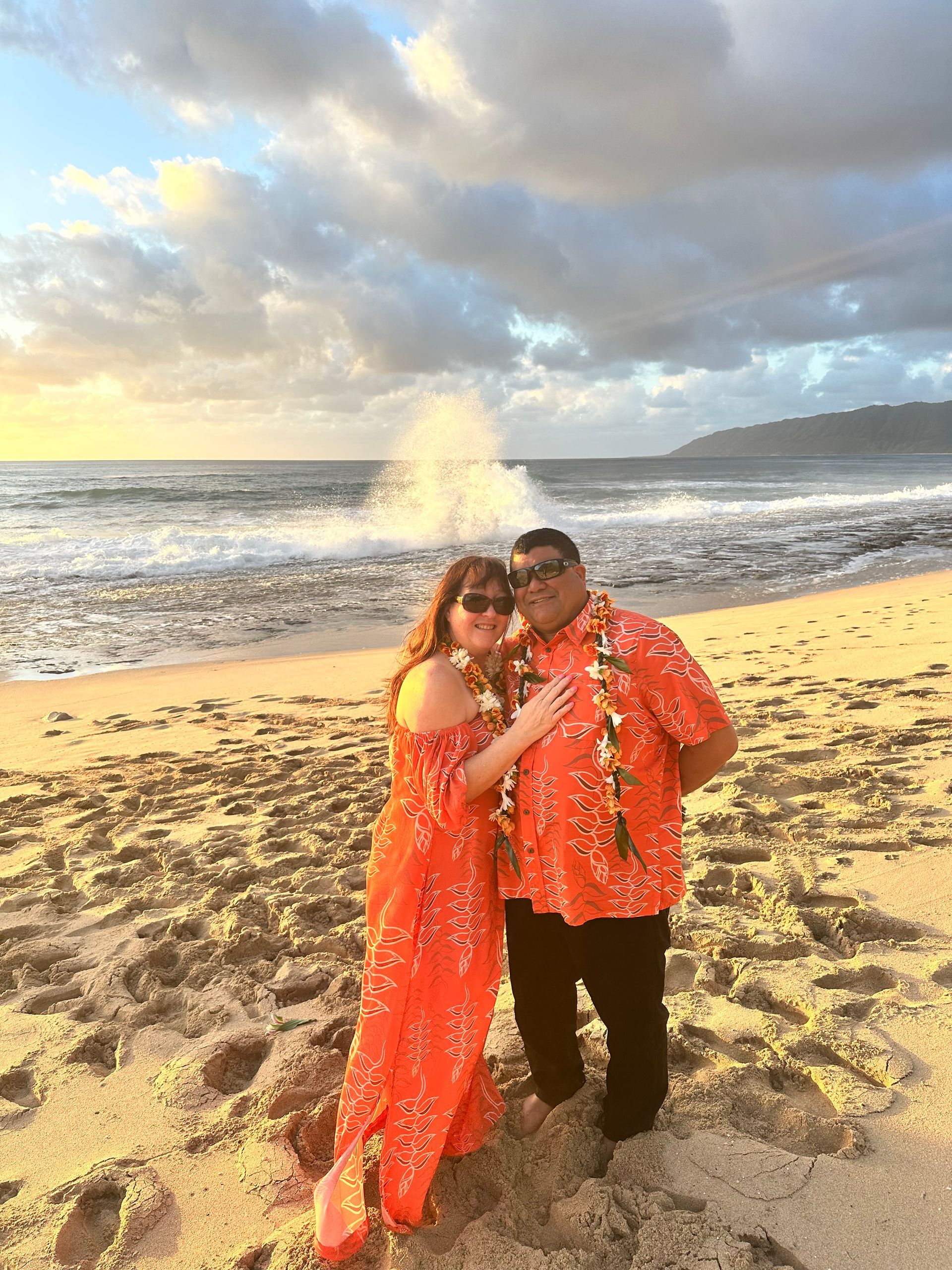 Couple on beach wearing orange floral attire, sunset, waves crashing.