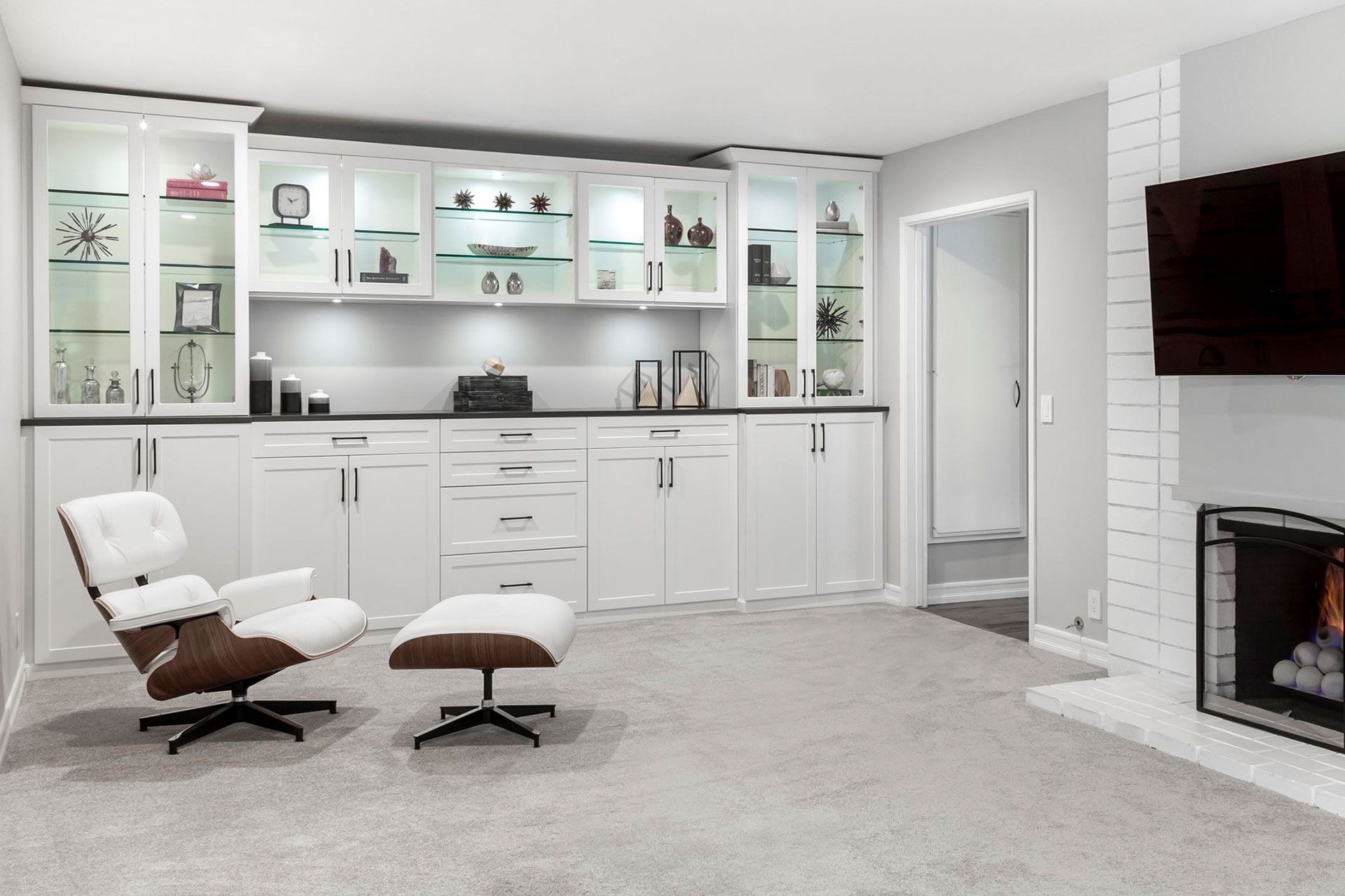 Modern white built-in cabinetry with glass display shelves, a fireplace, and a chair in a room with light grey carpet.