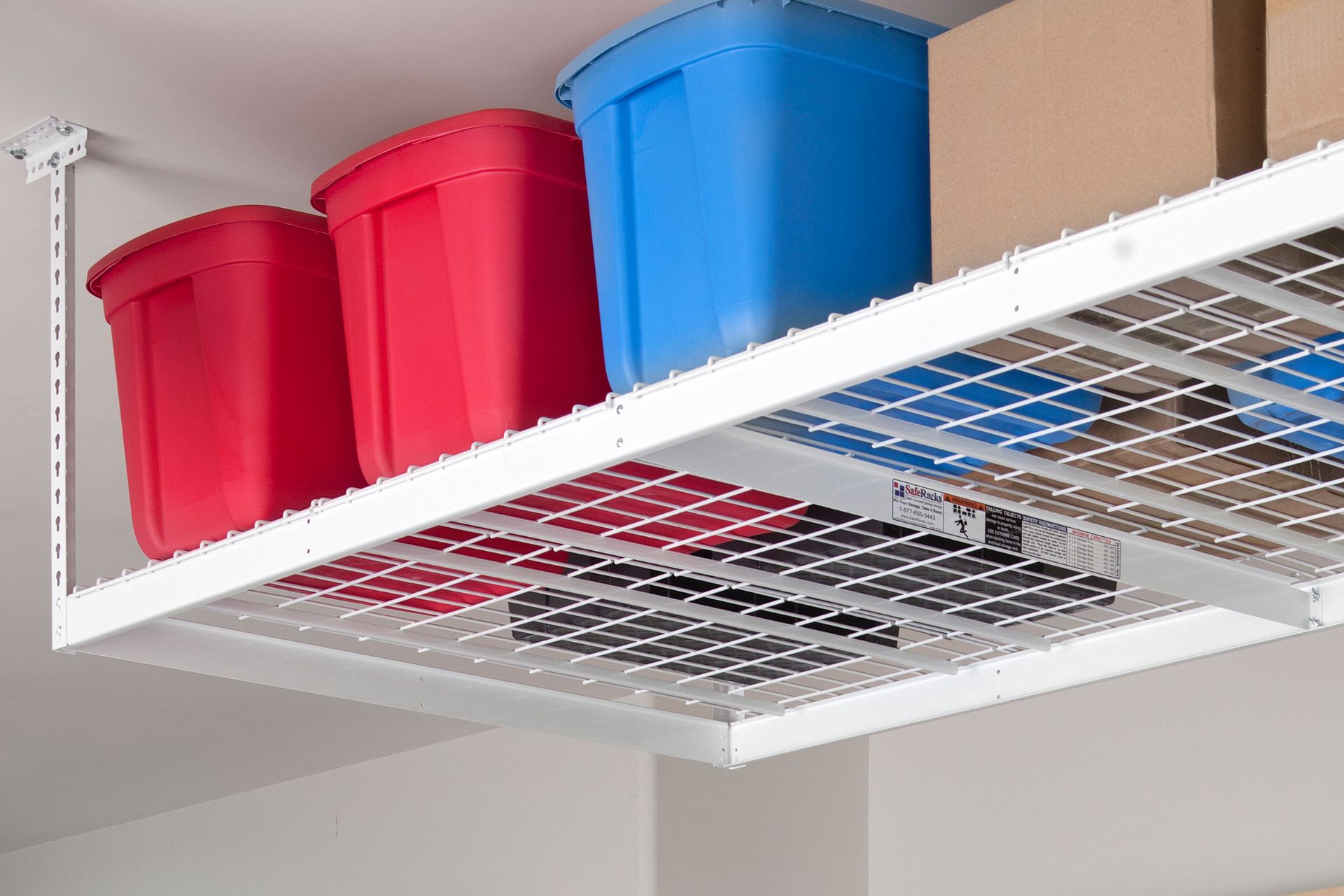 White metal overhead storage rack in a garage, holding red and blue storage bins, and cardboard boxes.