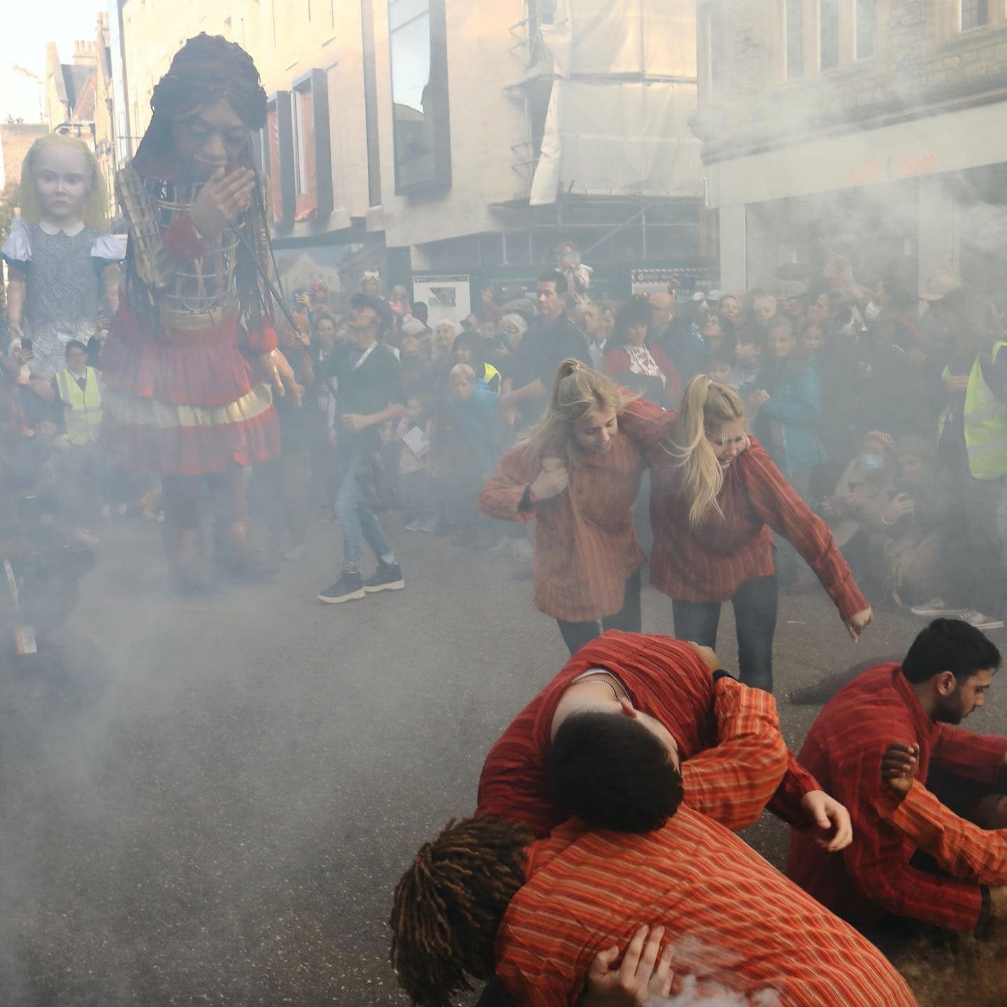 A large scale female puppet of a girl called Amal. She is from Syria. She is standing Next to another large scale Puppet of a girl with blonde hair called Alice.A group of people are  infant of them in couples they are holding each other.They recover from the ground. There is smoke. There are people standing around the action in  a crowd.