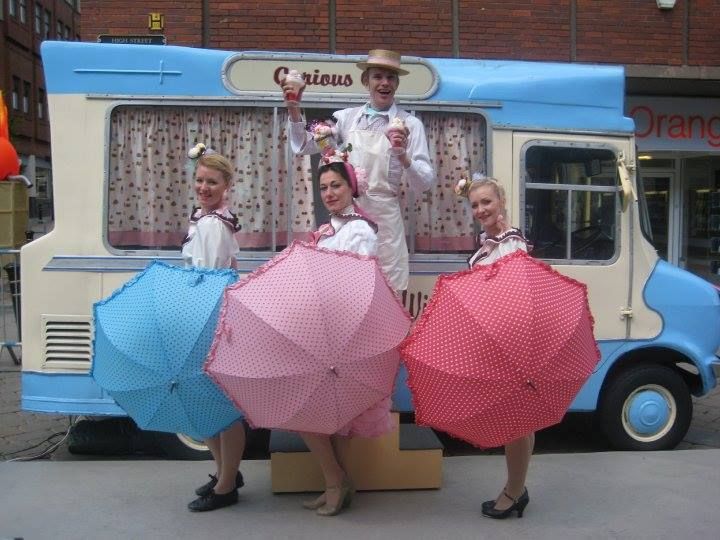 A group of  performers dressed in ice cream coloured vintage dress from the 1940s are standing in front of a blue and cream  vintage ice cream van. Three female dancers are holding polka dot umbrellas in red, blue and pink. The man is wearing a straw boater hat and is holding knickerbocker glory ice creams in his hands. They all look really happy
