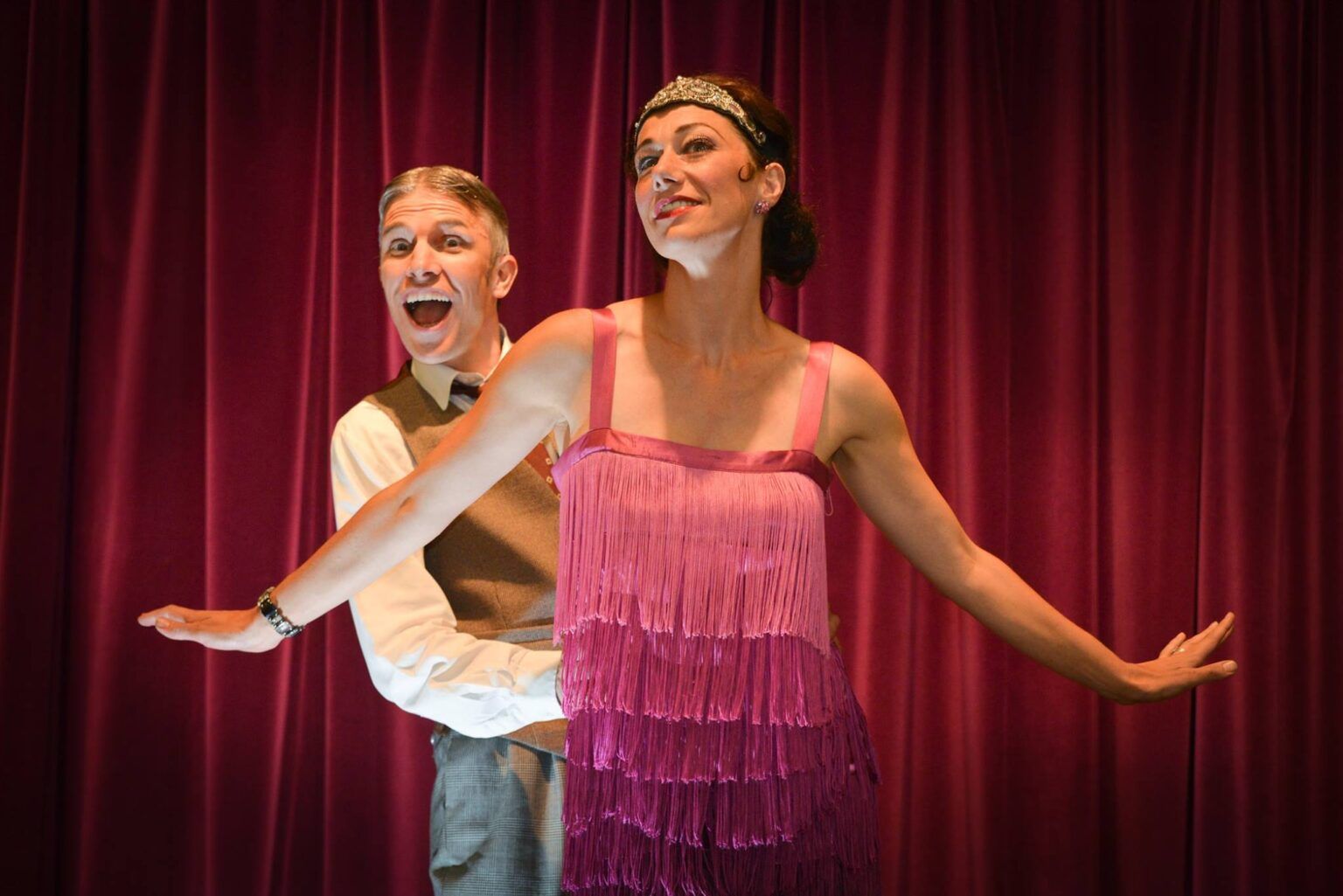 A man and a woman are dancing on a stage in front of a red curtain. The woman is wearing a pink 19202 fringed dress.