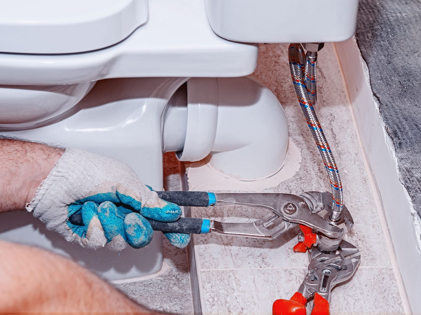 Gloved hands using pliers to repair plumbing under a sink, with exposed pipes and fittings