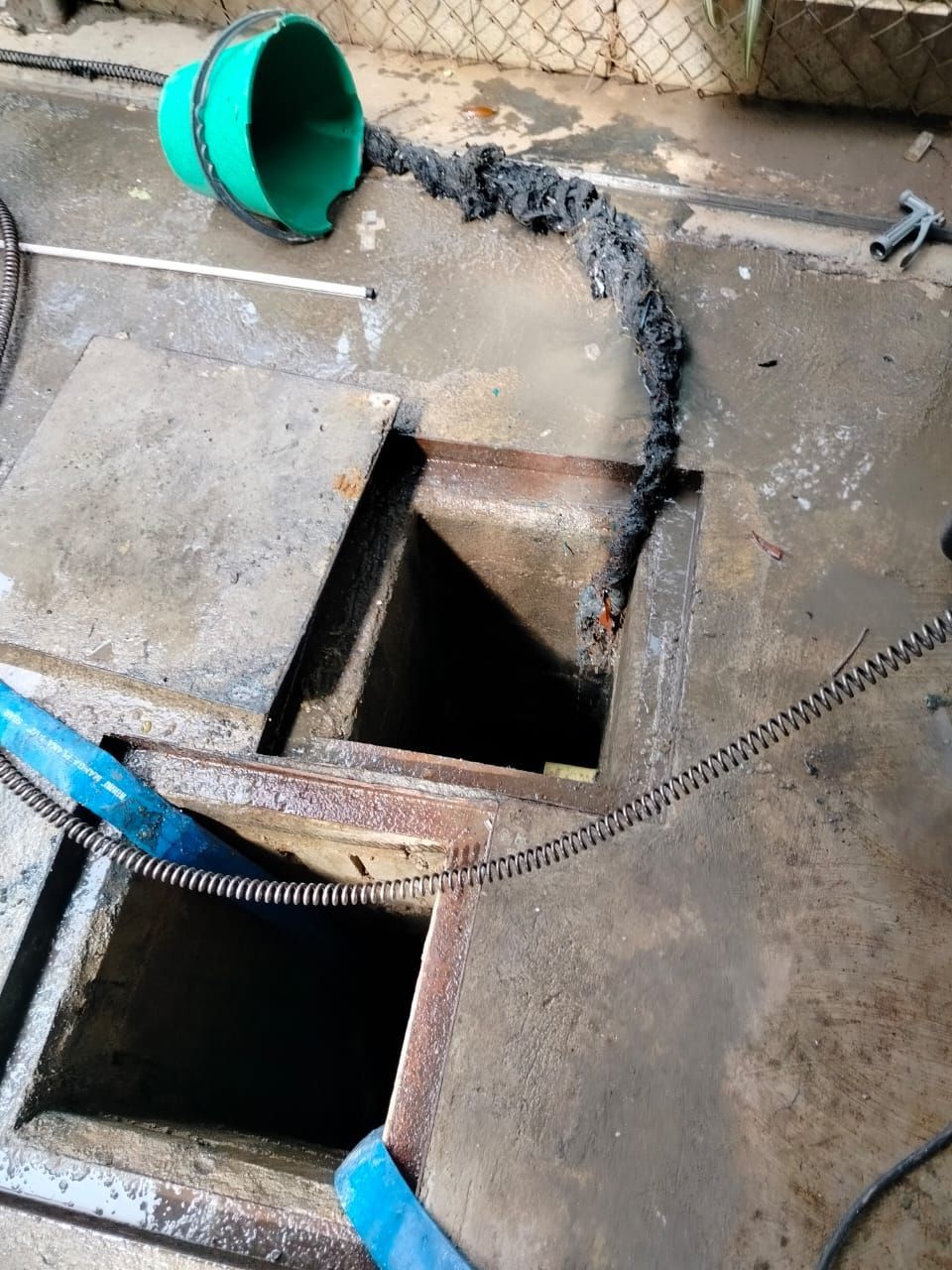 Pozo de registro abierto en suelo de hormigón con un cubo verde y una barrera de cadena cerca.