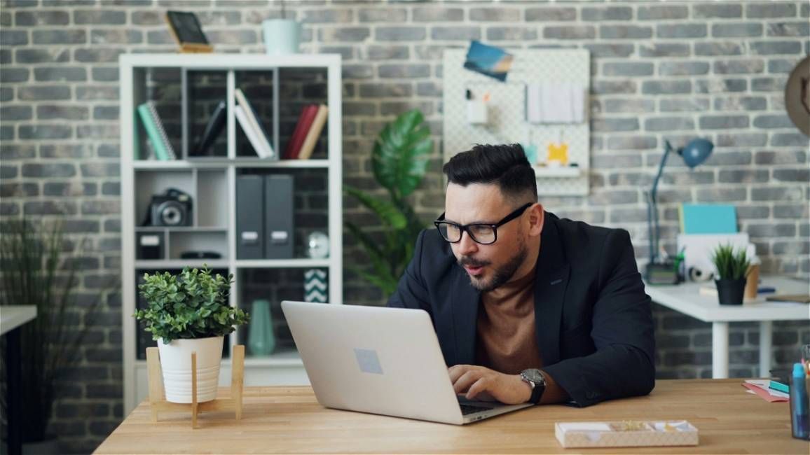A man with glasses in his office is looking at his laptop with excitement.