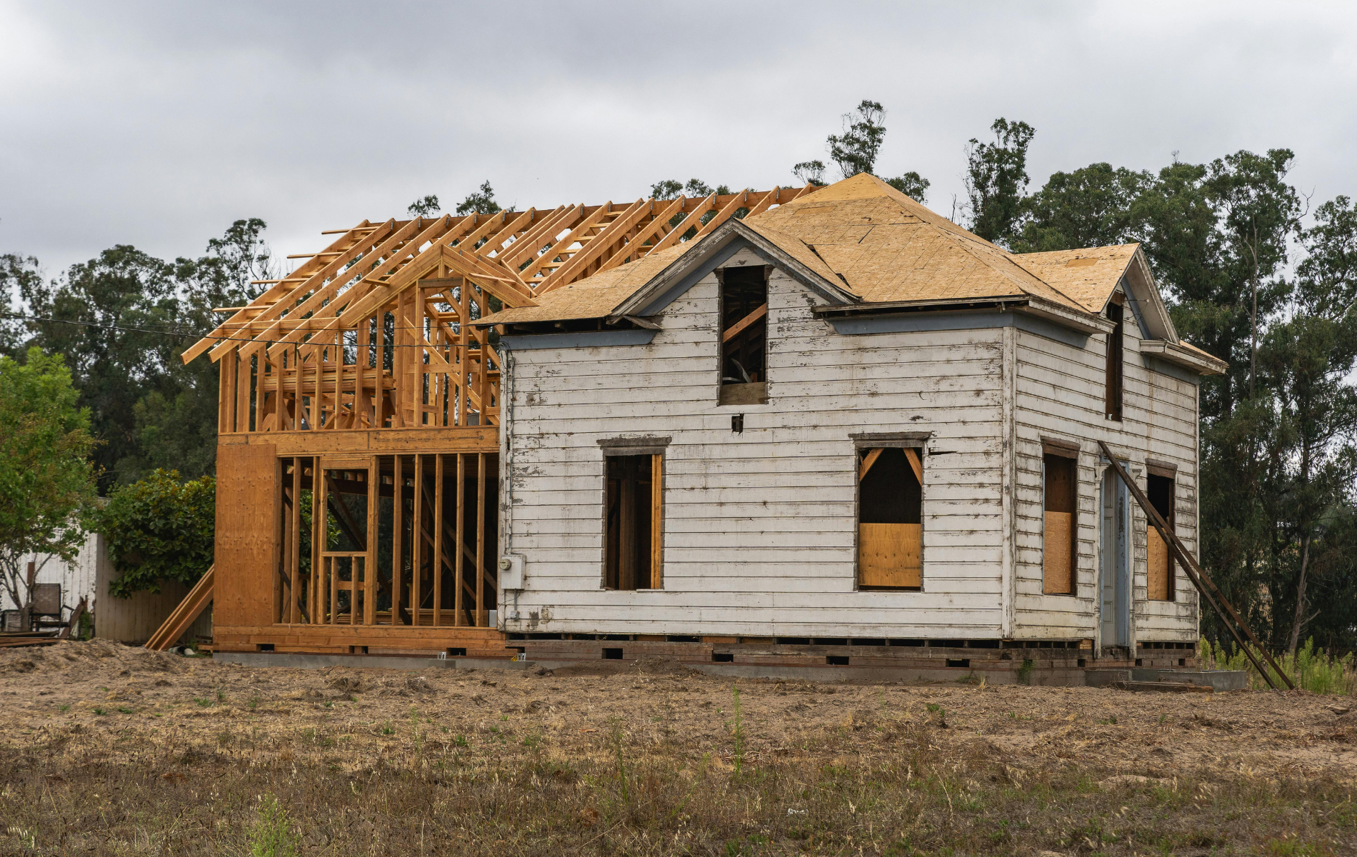 An old white wooden house under construction, featuring a newly framed addition on its left side.