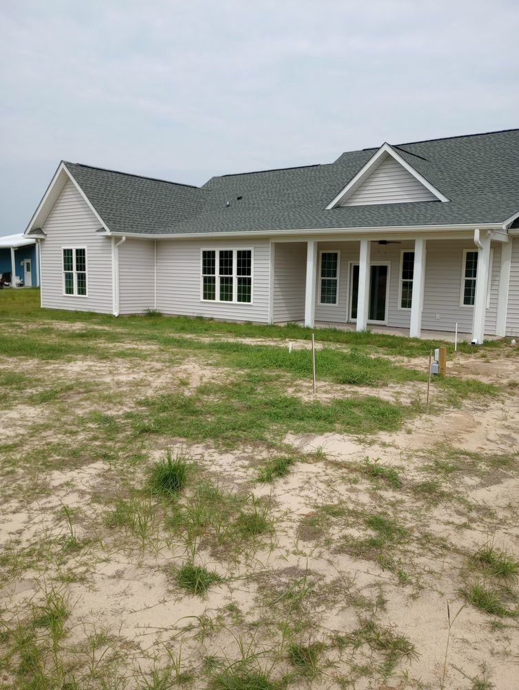 A newly constructed, single-story house with light siding and a grey shingled roof, surrounded by sandy, sparse grass.