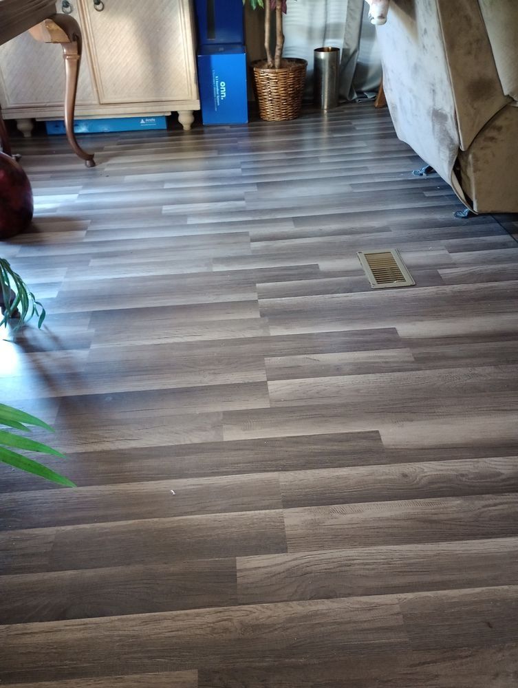 Gray-toned wood-look laminate flooring in a room with a floor vent, furniture legs, and a potted plant in the background.