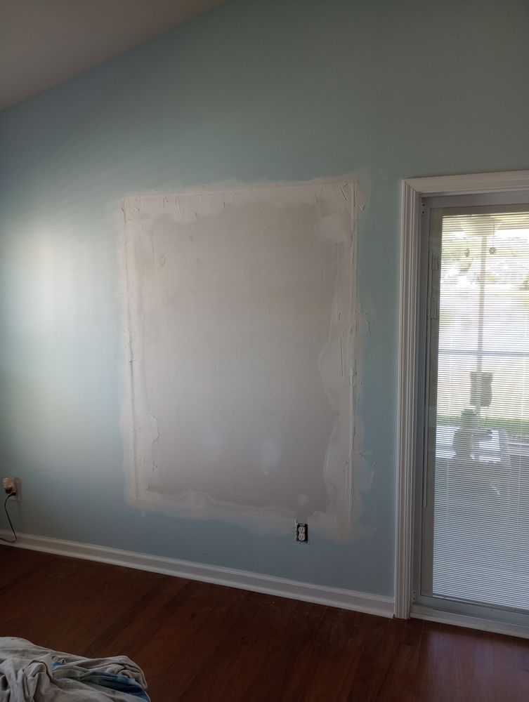 A light blue wall with a large, unpainted drywall patch in the center, near a sliding glass door and hardwood floors.