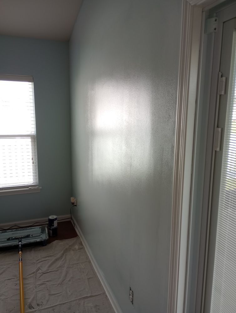 A room interior features a light blue wall being painted, with a paint roller on the floor and a window to the left.