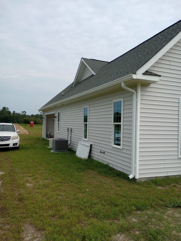 Side view of a light-colored, single-story house with dark shingles, two windows, and a white downspout on a grassy lot.